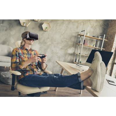 Eine Person sitzt entspannt mit den Füßen auf dem Tisch, trägt eine VR-Brille und hält einen Spielcontroller.