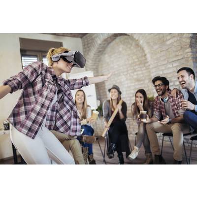 Eine Frau trägt ein VR-Headset und bewegt sich, während fünf lachende Personen zuschauen. Alle sitzen entspannt in einem gemütlichen Raum.