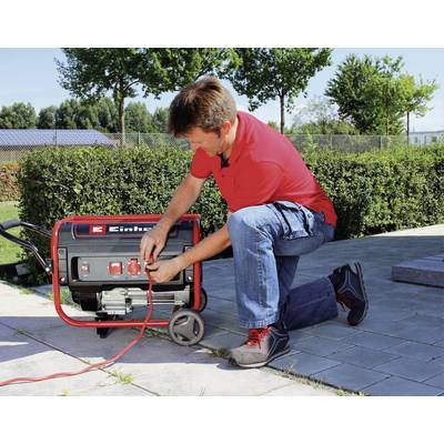 Ein Mann kniet und bedient ein rot-schwarzes Elektroaggregat im Freien, umgeben von Pflanzen und einem gepflasterten Weg.