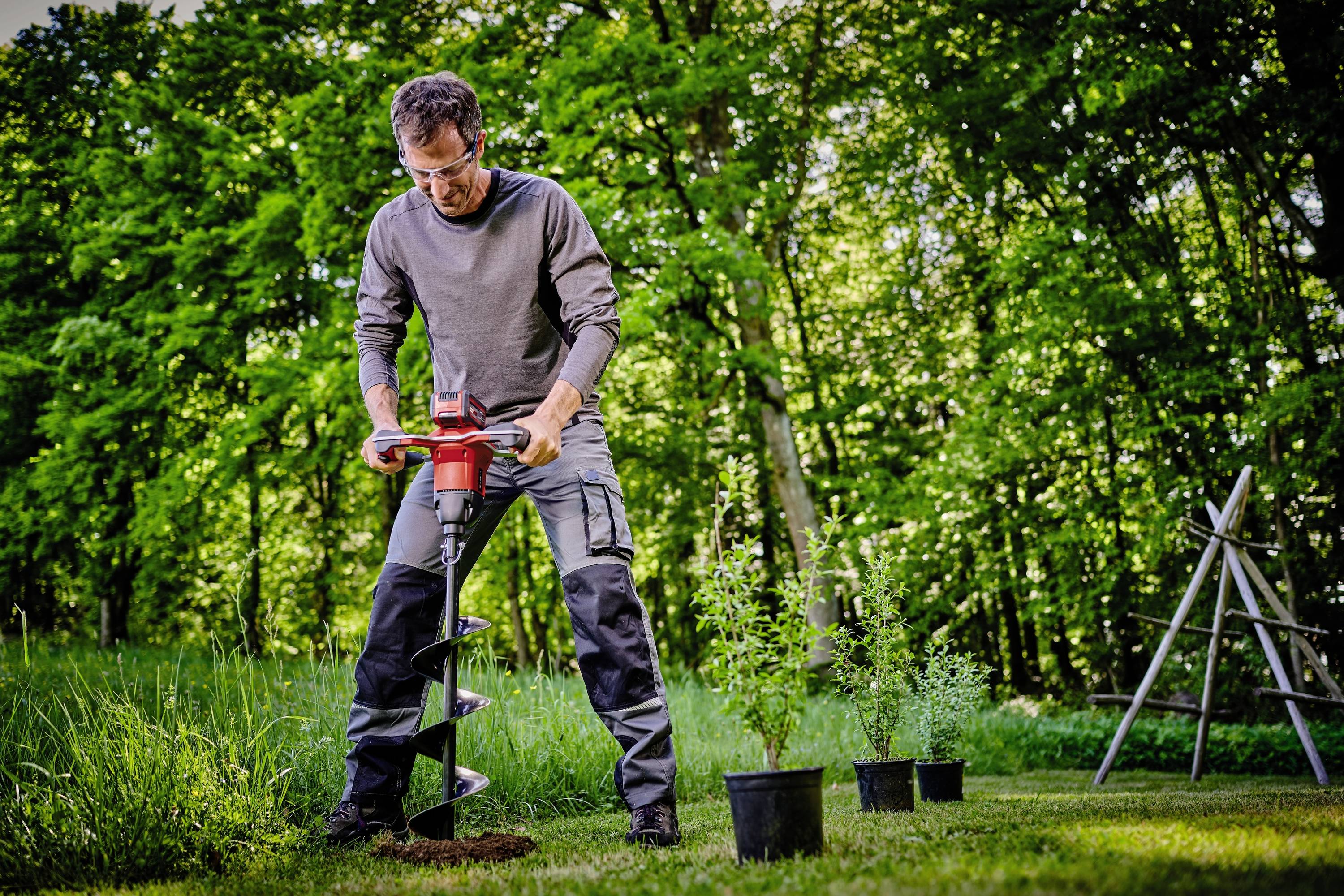 Person bohrt mit einer Erdbohrmaschine im Garten, umgeben von Pflanzen in Töpfen und einem Baum im Hintergrund.