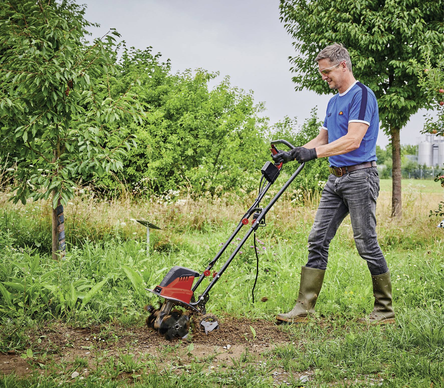 Ein Mann mit blauer Kleidung und Gummistiefeln bedient einen motorisierten Gartenfräse, um den Boden in einem grünen Garten umzugraben.