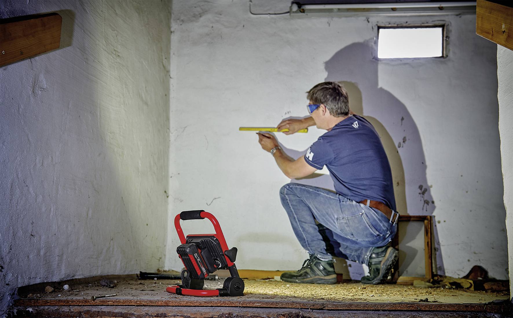 Ein Mann im T-Shirt und Jeans kniet in einem weiß gestrichenen Raum und misst mit einem Maßband. Eine Arbeitsleuchte steht am Boden.