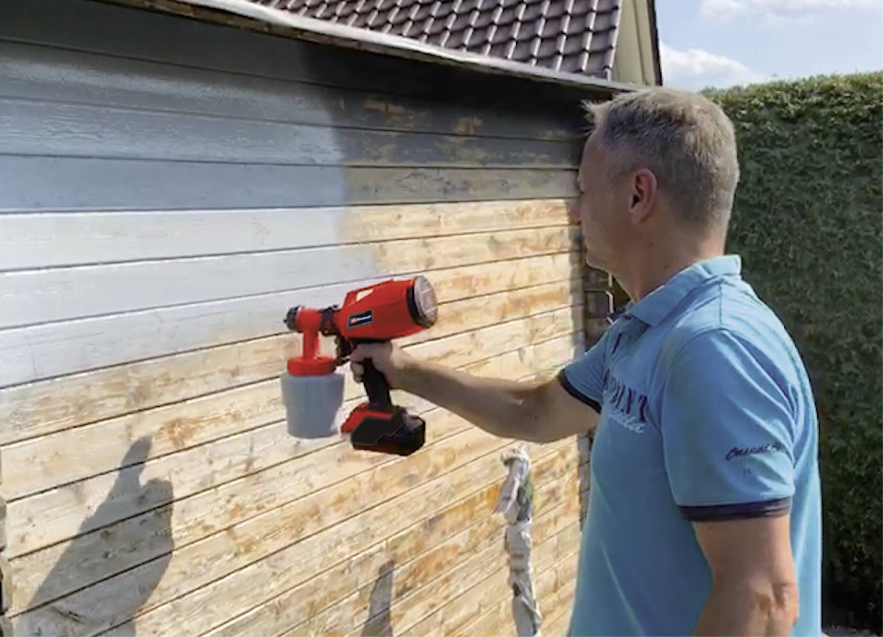 Mann sprüht dunkle Farbe mit einer Farbspritzpistole auf eine Holzwand; die Wand ist teilweise gestrichen.