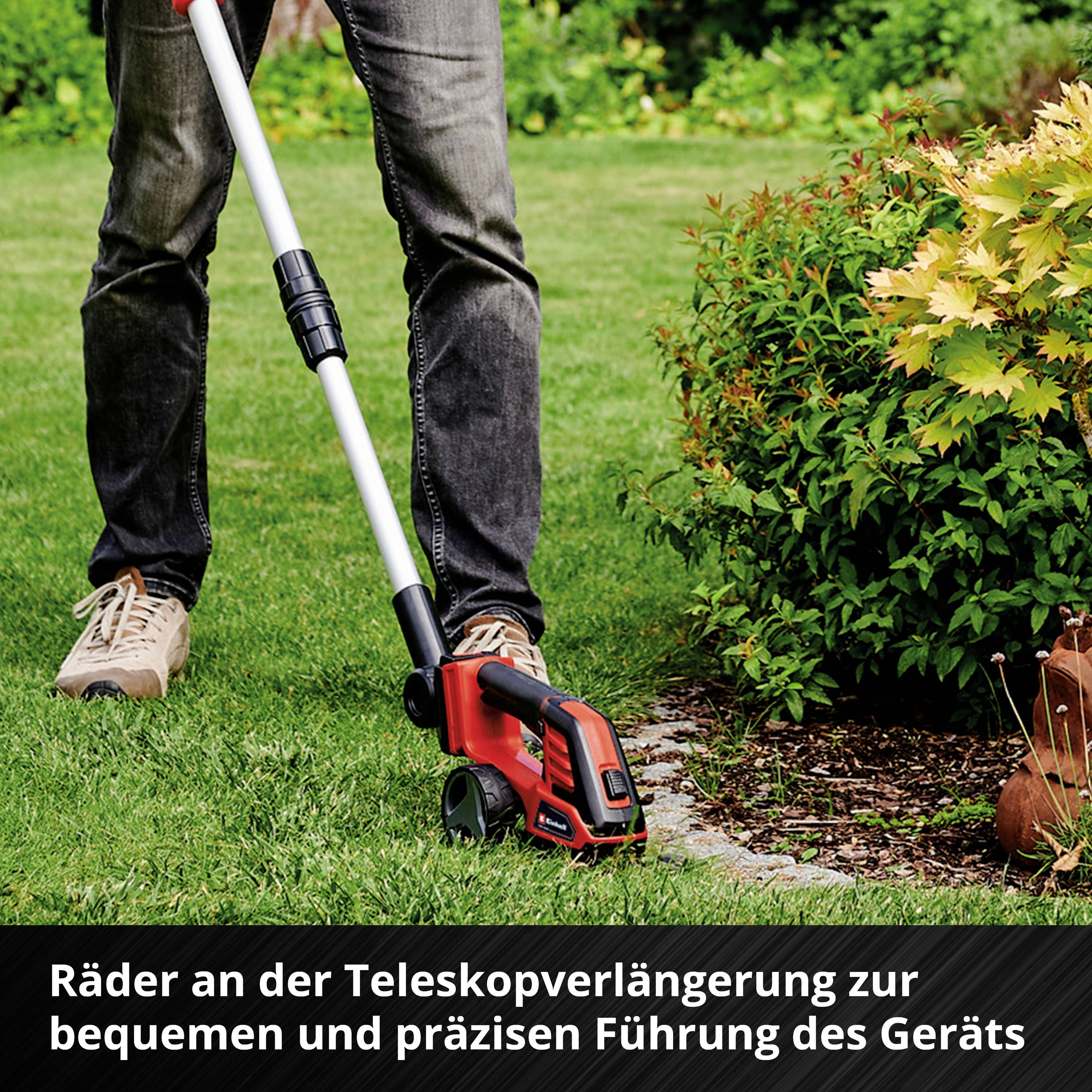 Person mäht den Rasen mit einem elektrischen Trimmer. Werkzeug auf Teleskopverlängerung; daneben eine blühende Pflanze.