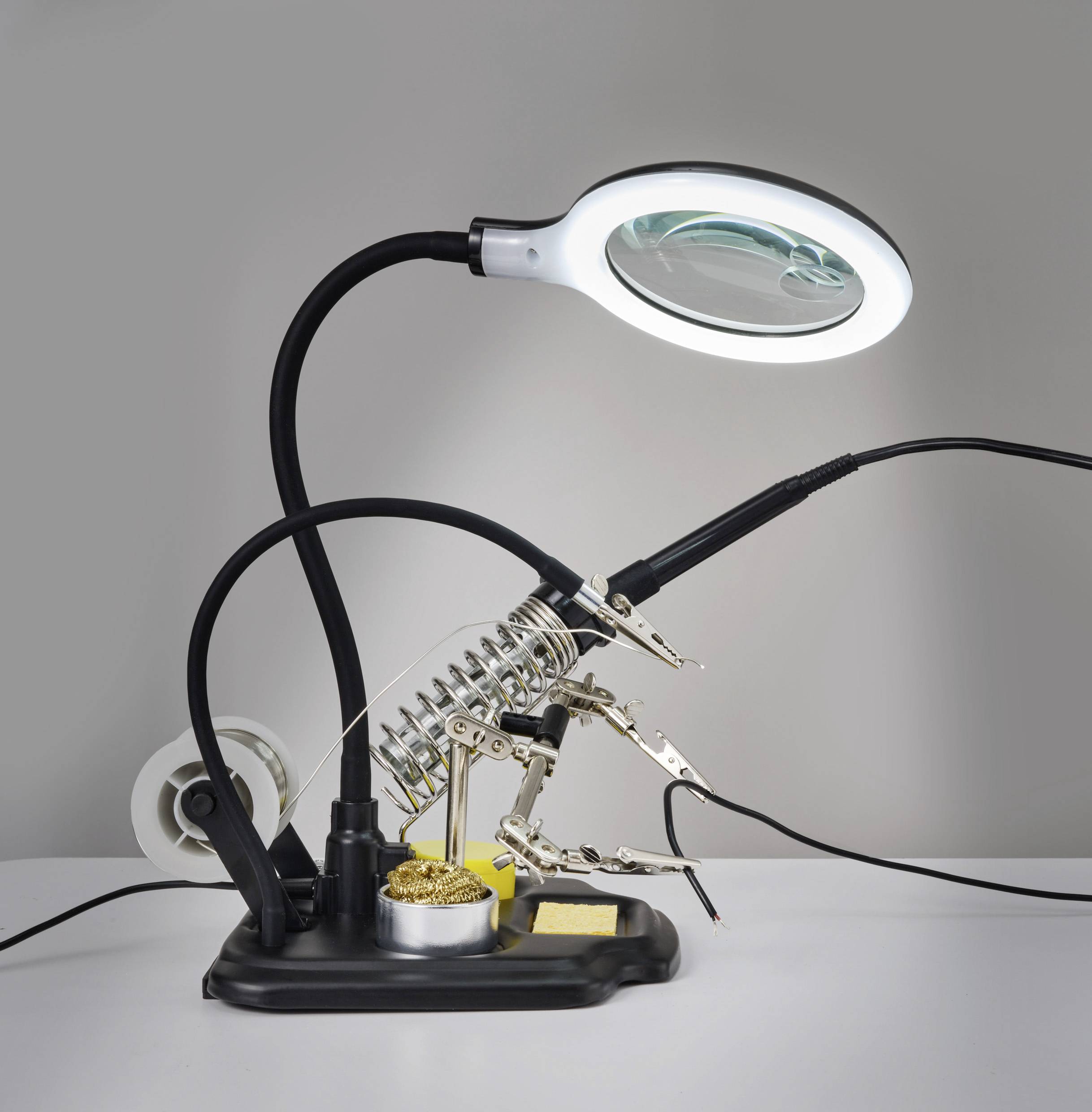 Eine Lupe mit verstellbarem Arm ist beleuchtet und über einer Lötstation positioniert, die mit Werkzeughaltern und Lödzubehör ausgestattet ist.