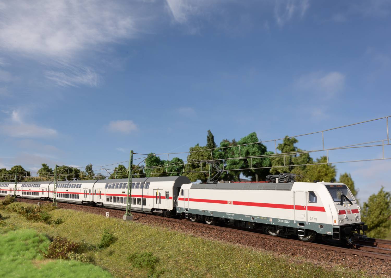 Ein weißer Zug mit roten Streifen fährt auf einer elektrifizierten Strecke durch eine grüne Landschaft unter blauem Himmel.