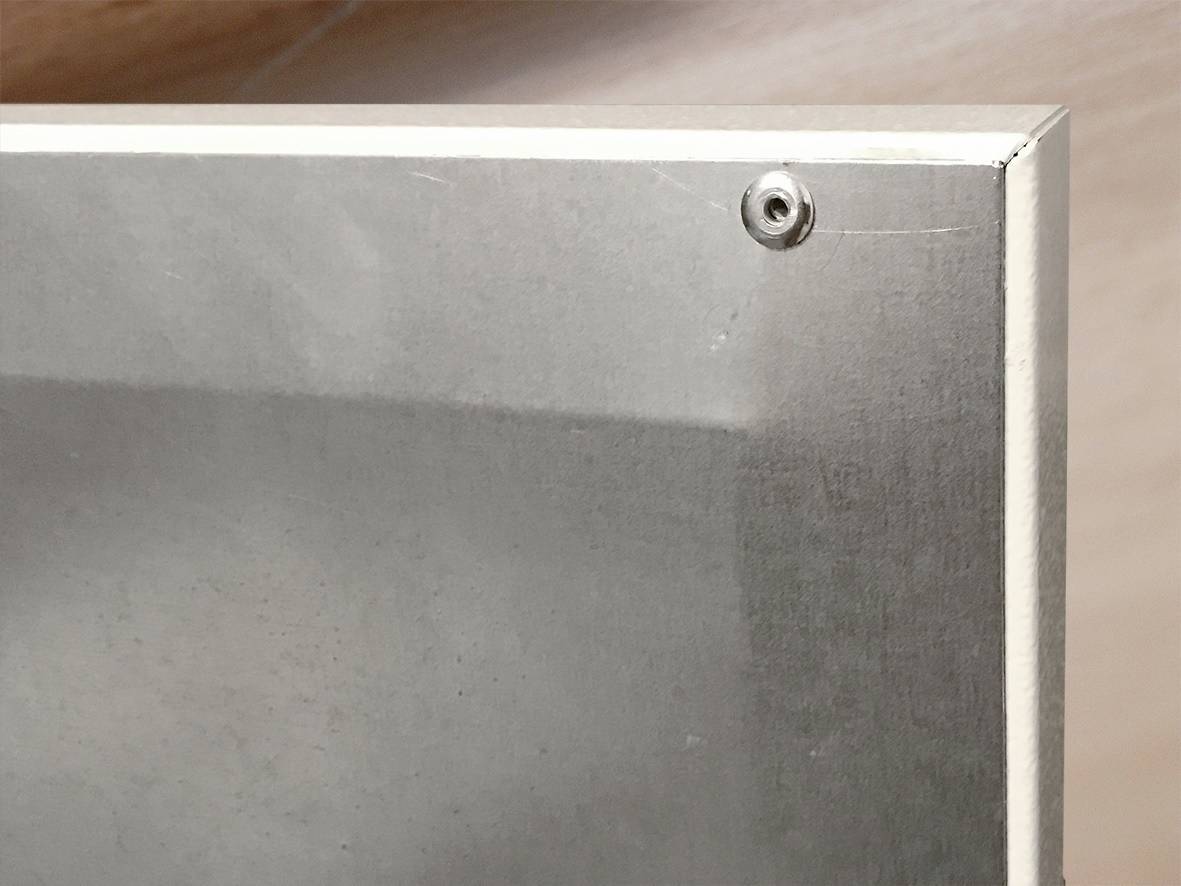 Ecke eines Metallvierecks, befestigt mit einer Schraube in der oberen rechten Ecke. Matt und glänzend, ohne sichtbare Gebrauchsspuren.