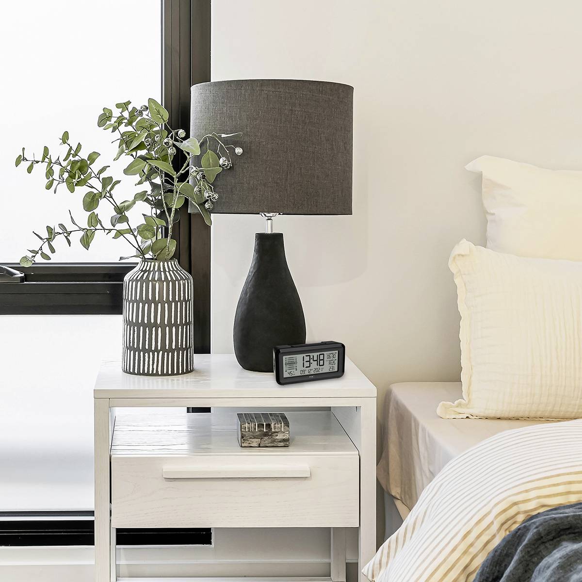 Nachttisch mit Tischlampe, Wecker zeigt 3:40 an, Pflanze in gestreifter Vase. Schlafzimmerumgebung mit neutralen Farben.