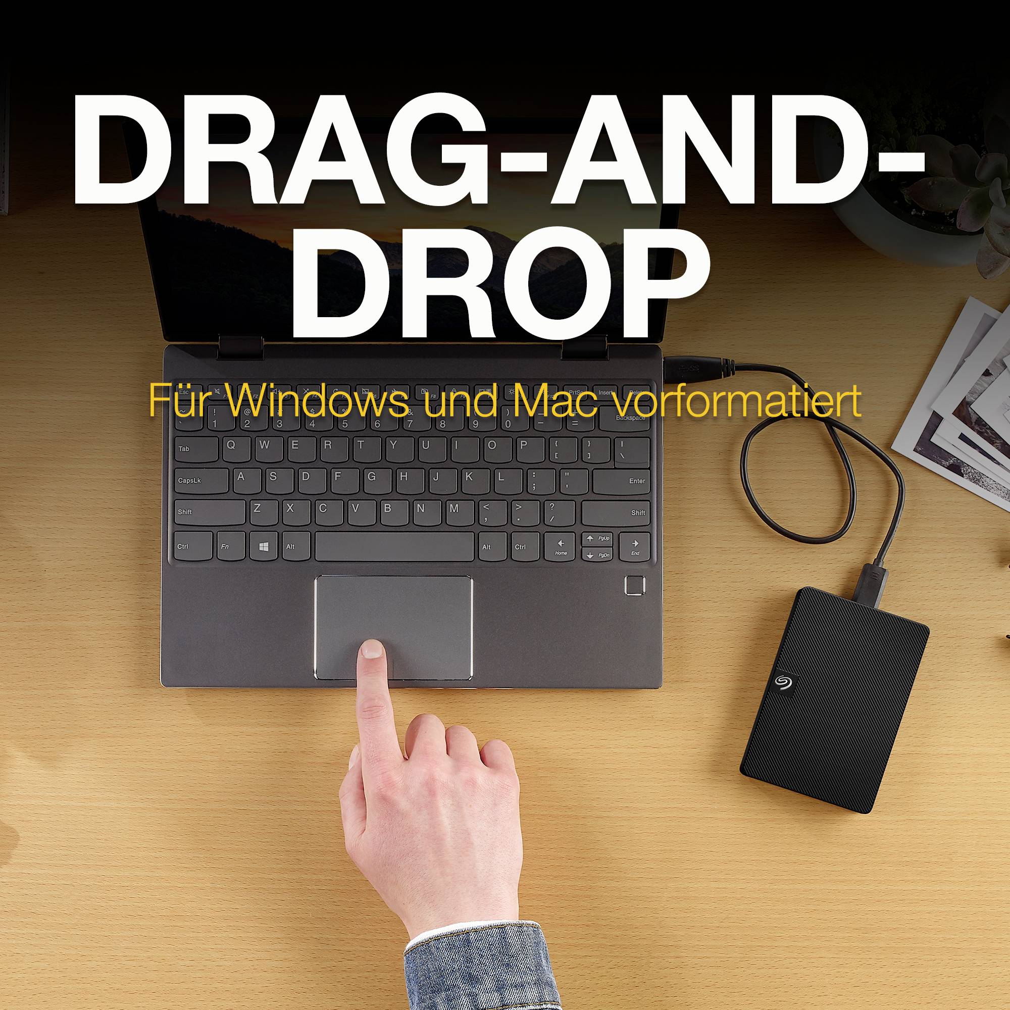 Eine Hand hält ein schwarzes externes Laufwerk neben einem Laptop auf einem Schreibtisch. Text auf dem Bild: 'Drag-and-Drop, für Windows und Mac vorformatiert'.