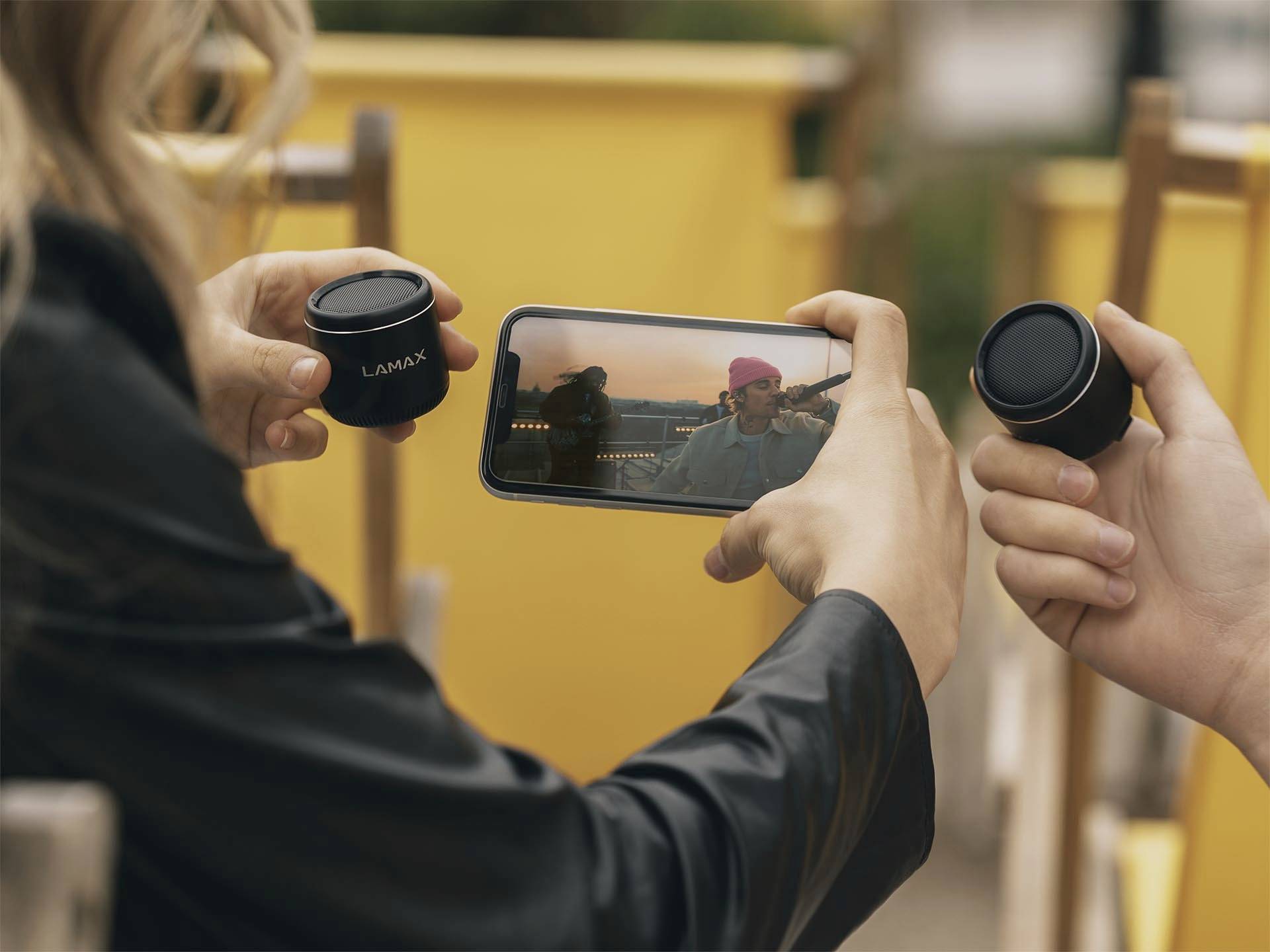 Eine Person hält ein Smartphone, auf dem ein Video abgespielt wird, während zwei Hände würfelförmige Lautsprecher in der Nähe halten.