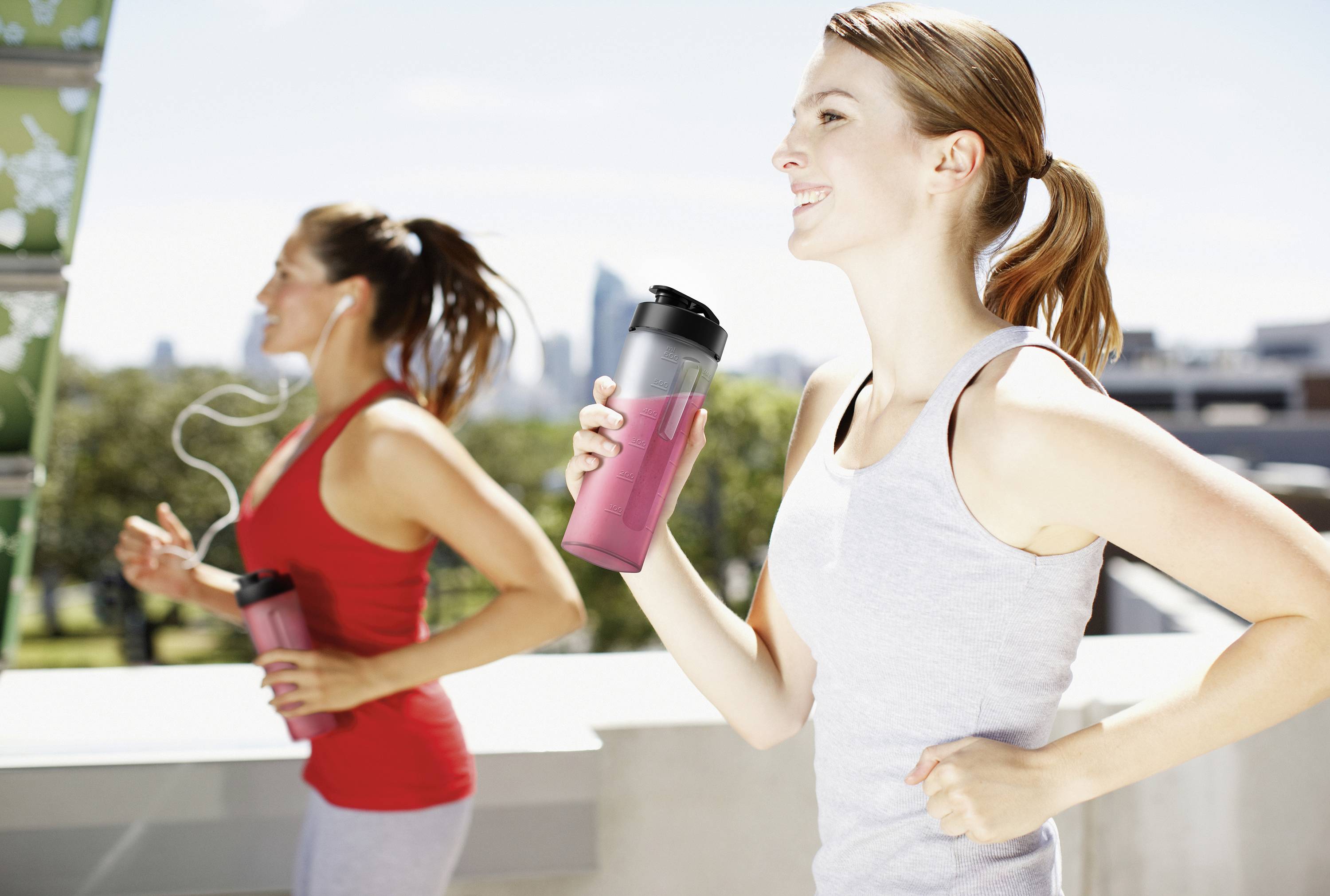 Zwei Frauen joggen im Freien, halten Trinkflaschen in der Hand, tragen sportliche Kleidung, im Hintergrund unscharfe Stadtansicht.