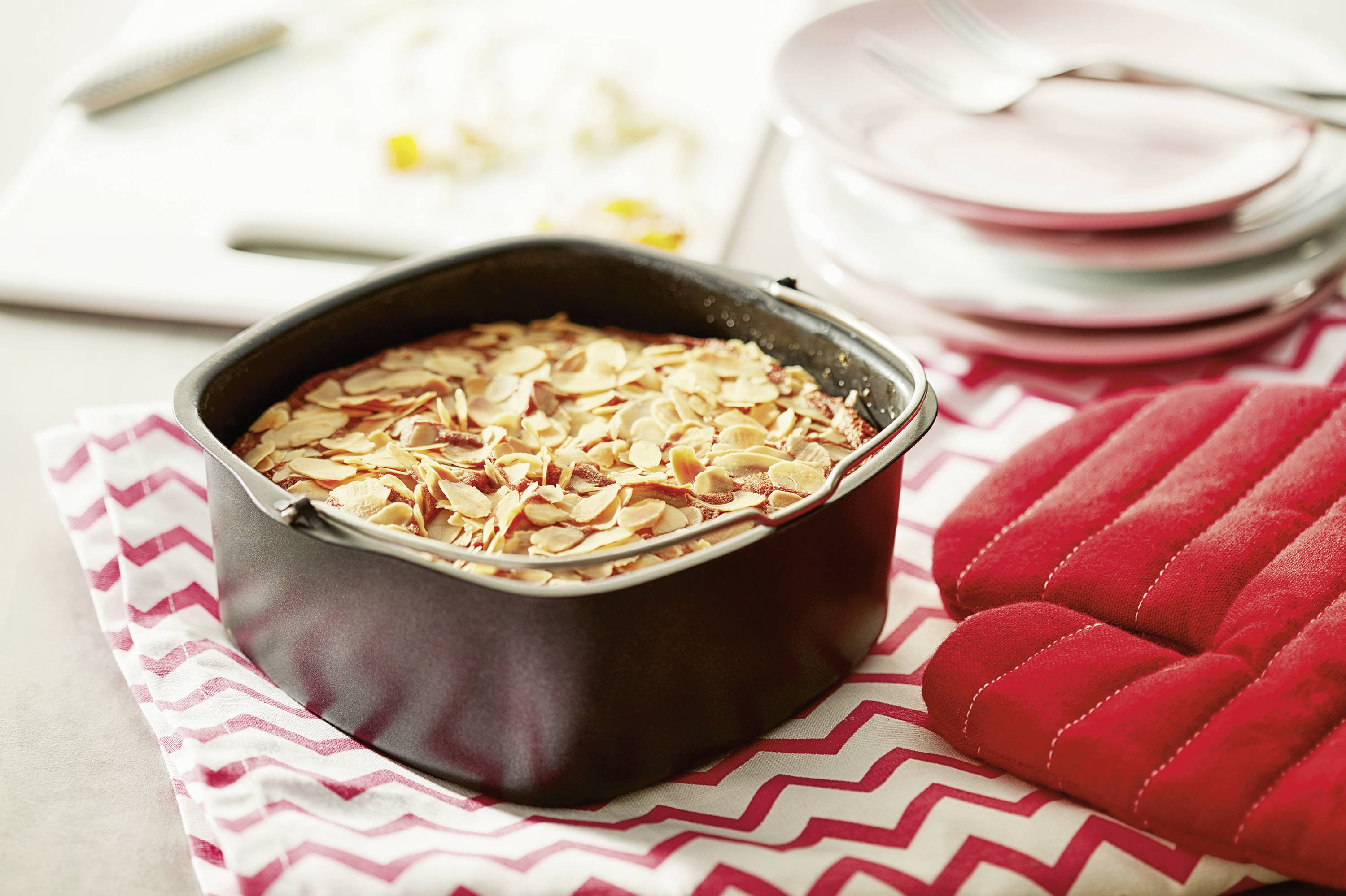 Ein frisch gebackener Mandelkuchen in einer schwarzen Backform steht auf einem Tisch mit rot-weißem Zickzack-Muster und roten Ofenhandschuhen.