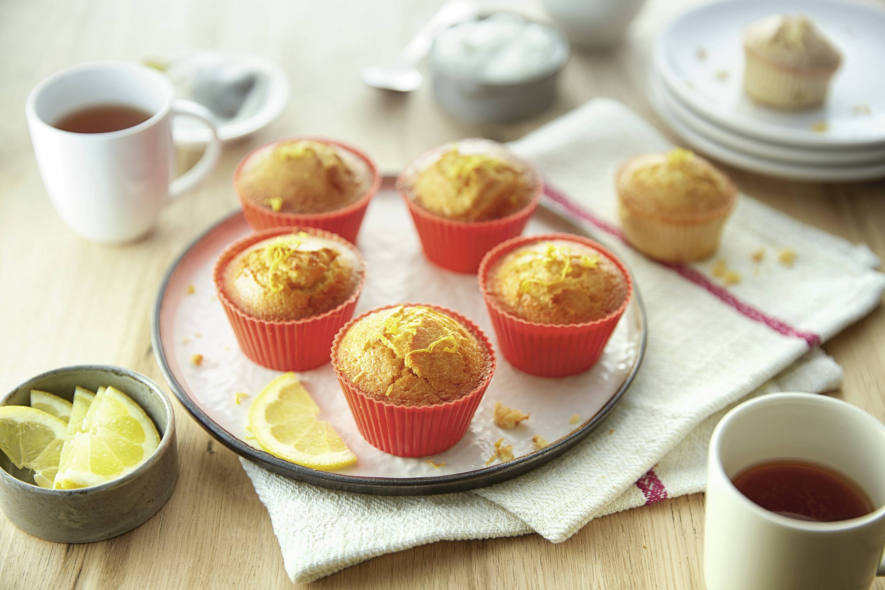 Auf einem Tisch stehen sechs Orangenmuffins in roten Förmchen auf einem Teller, umgeben von Zitronenscheiben und Tassen Tee.