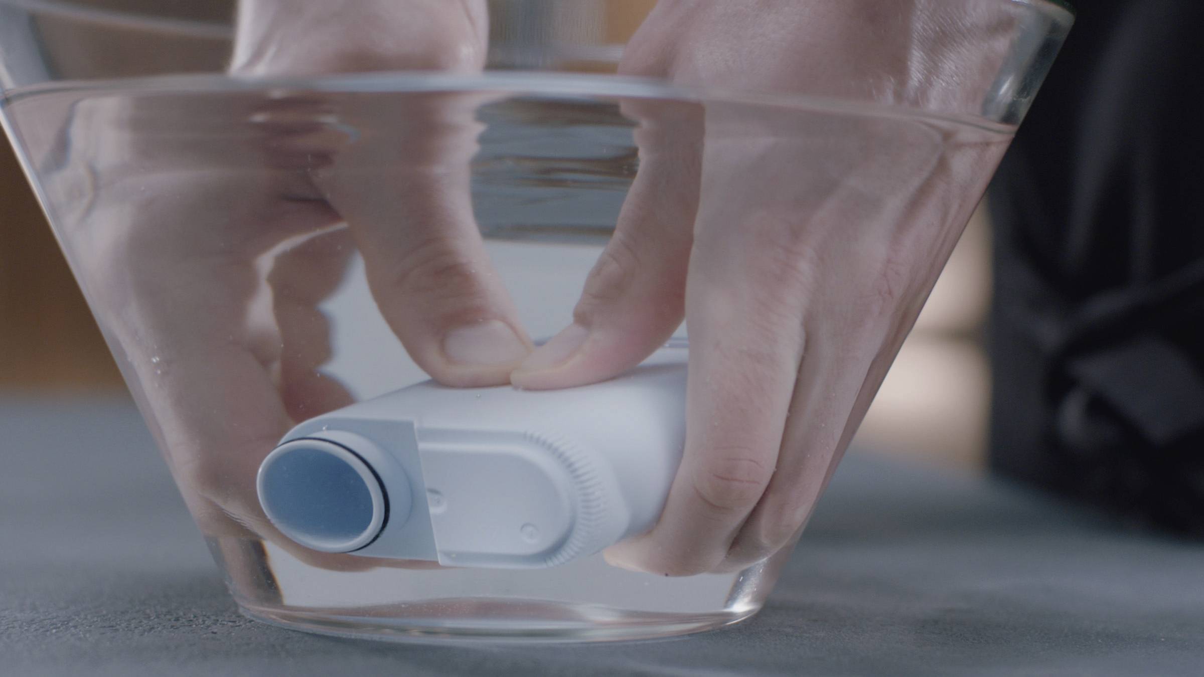 Hände tauchen eine kleine weiße Flasche mit blauem Deckel in eine Glasschüssel mit Wasser ein.