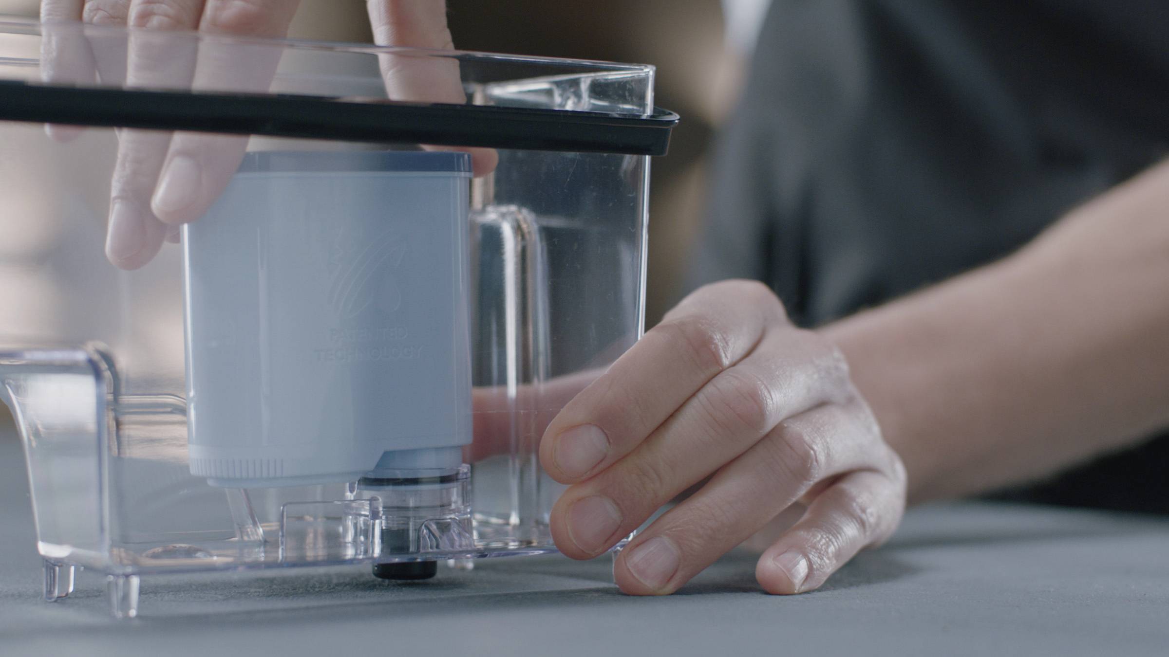 Person installiert einen blauen Wasserfilter in einem transparenten Behälter auf einem Tisch. Hände halten den Behälter vorsichtig.