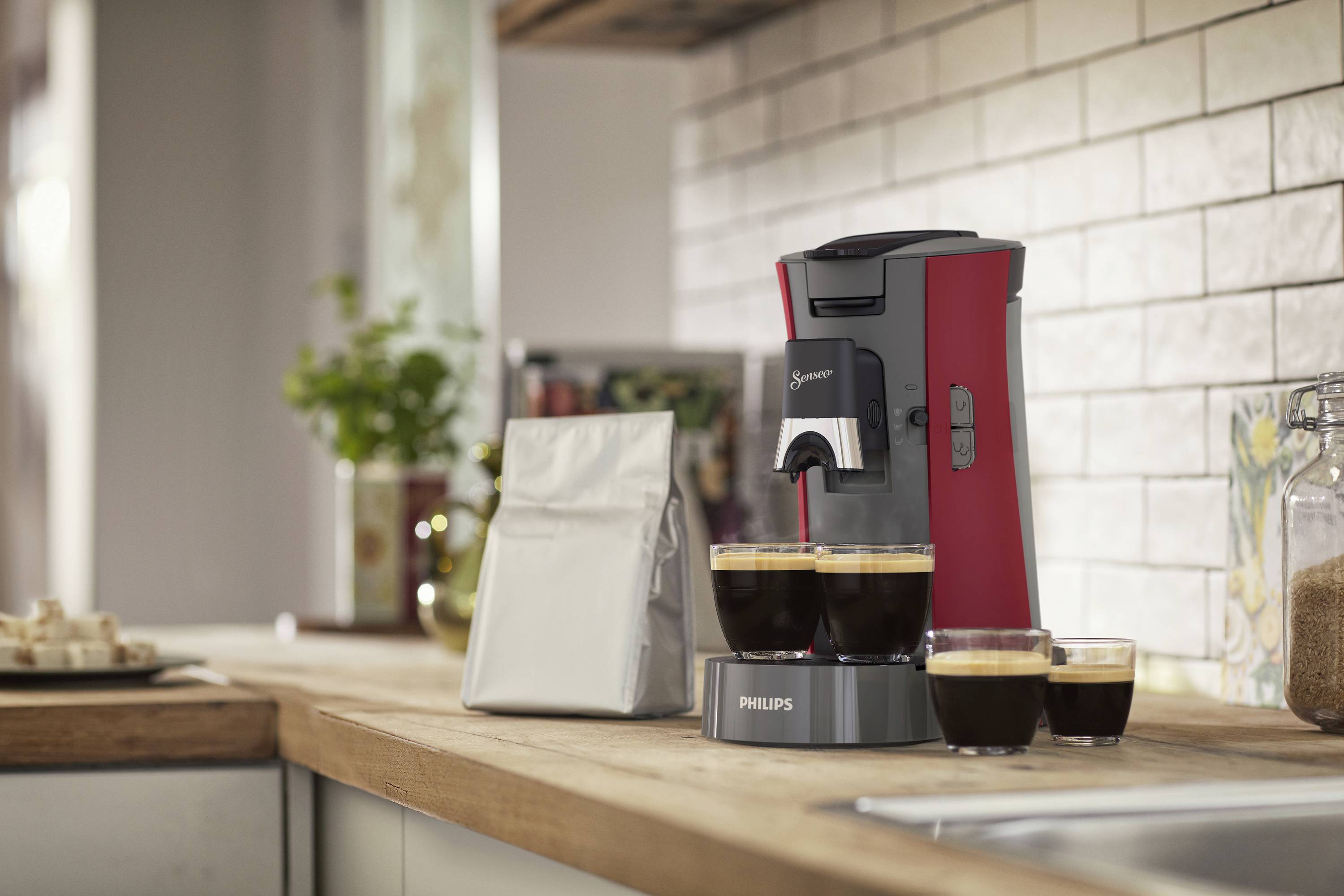 Rote Kaffeemaschine auf einer Küchenarbeitsplatte mit zwei gefüllten Tassen und einer Kaffeepackung daneben.