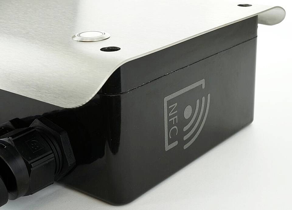 Ein schwarzes Gerät mit NFC-Symbol und einer silbernen Metallabdeckung. Links ist ein Kabelanschluss zu sehen.