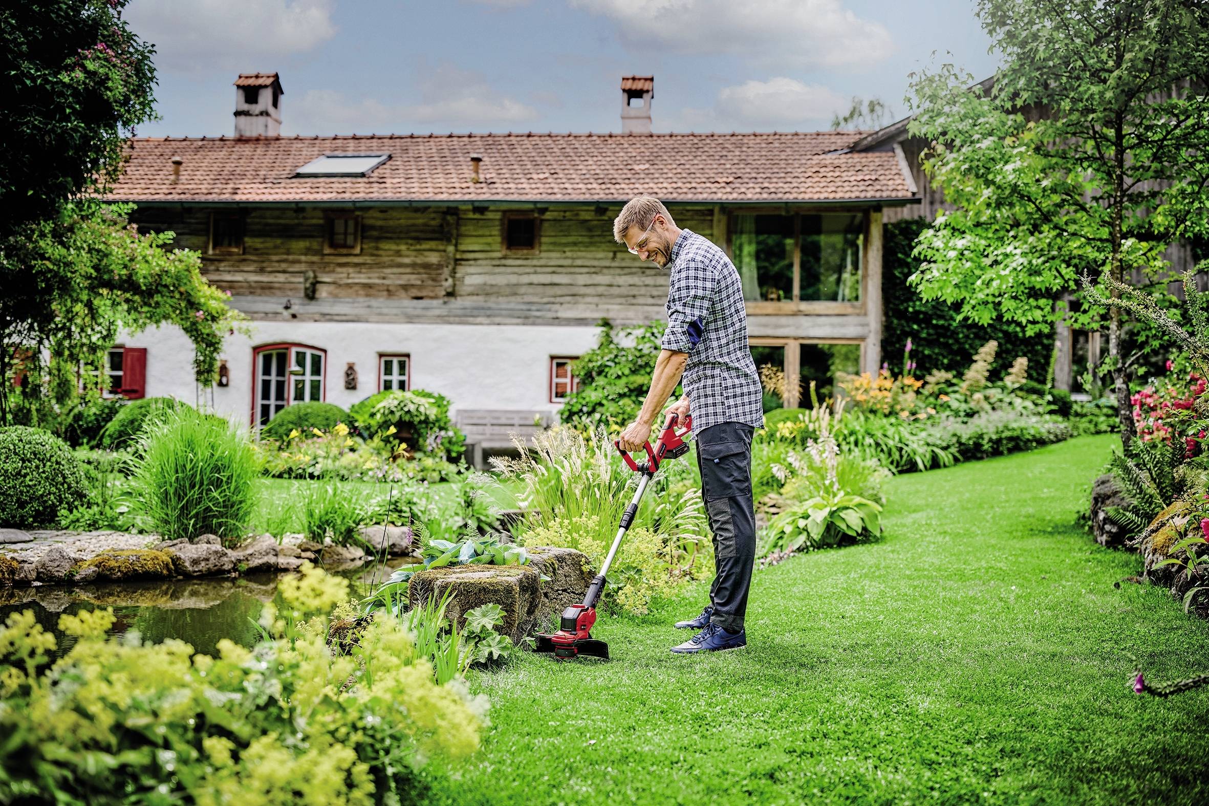 Ein Mann pflegt im Garten eines traditionellen Landhauses den Rasen. Die Umgebung ist grün und blühend.