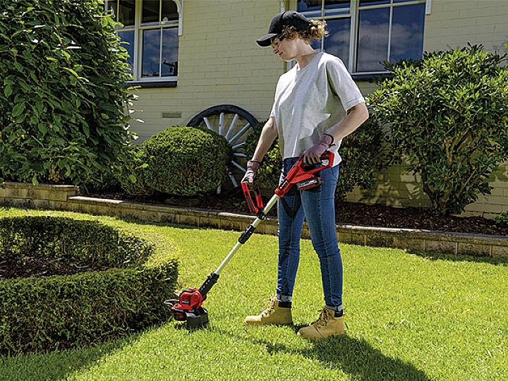 Eine Person trimmt Rasen mit einem elektrischen Rasentrimmer im Garten. Im Hintergrund sind Büsche und ein Gebäude zu sehen.