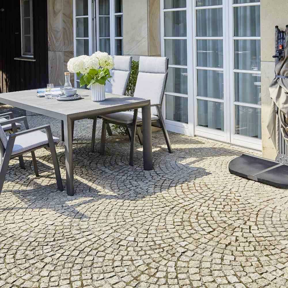 Terrasse mit rundem Pflastersteinboden, Tisch mit Blumenvase und Stühlen, Glas mit Wasser, Fensterfront im Hintergrund.