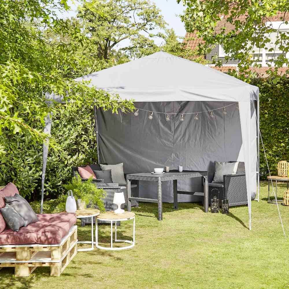Garten mit Pavillon, darunter ein Tisch und Stühle. Links im Bild eine Sitzgelegenheit auf Holzpaletten mit Kissen. Sonniger Tag.