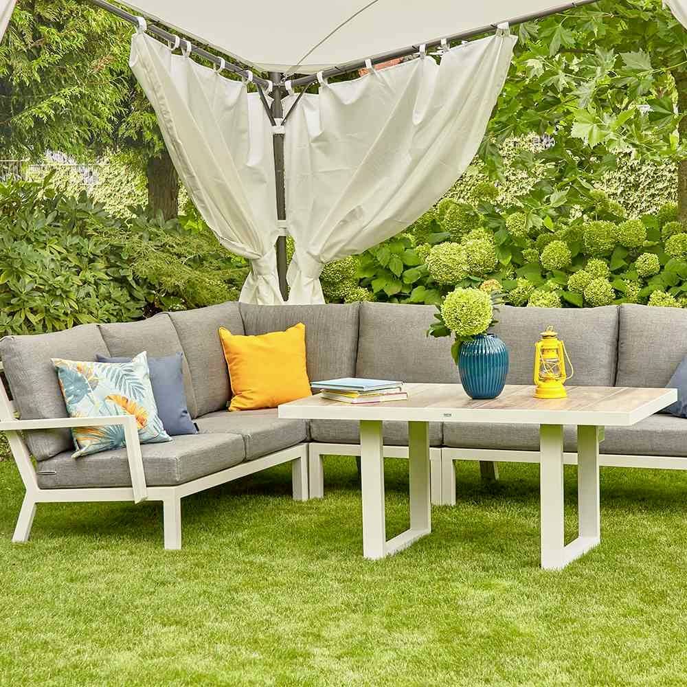 Grau-weiße Gartenlounge mit Kissen, Tisch und Pflanzen im Hintergrund.