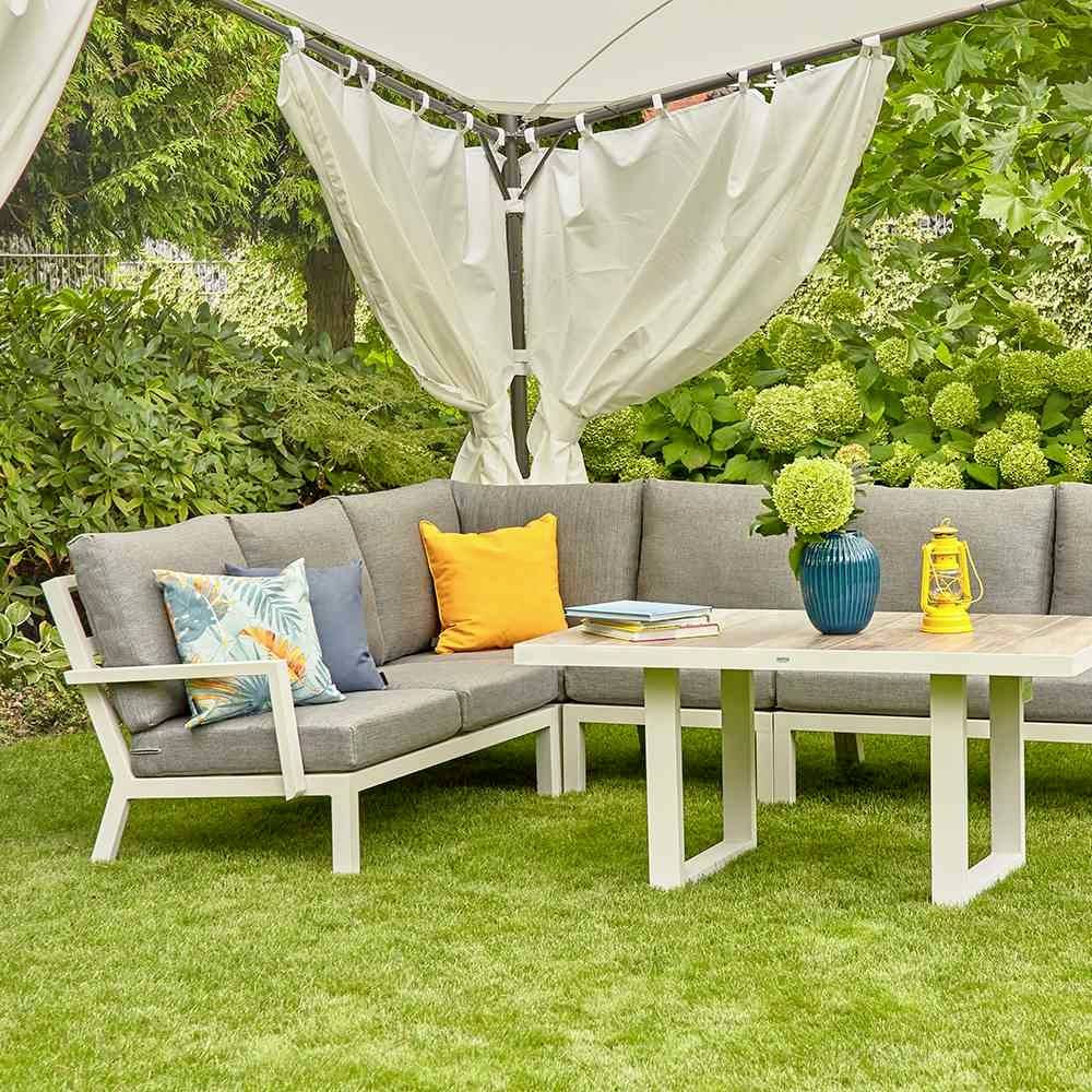 Ein Garten mit einer grauen Eck-Sofagarnitur und farbigen Kissen. Ein Holztisch steht vor dem Sofa mit einem blauen Krug und einem Buch darauf. Im Hintergrund sind grüne Pflanzen zu sehen.