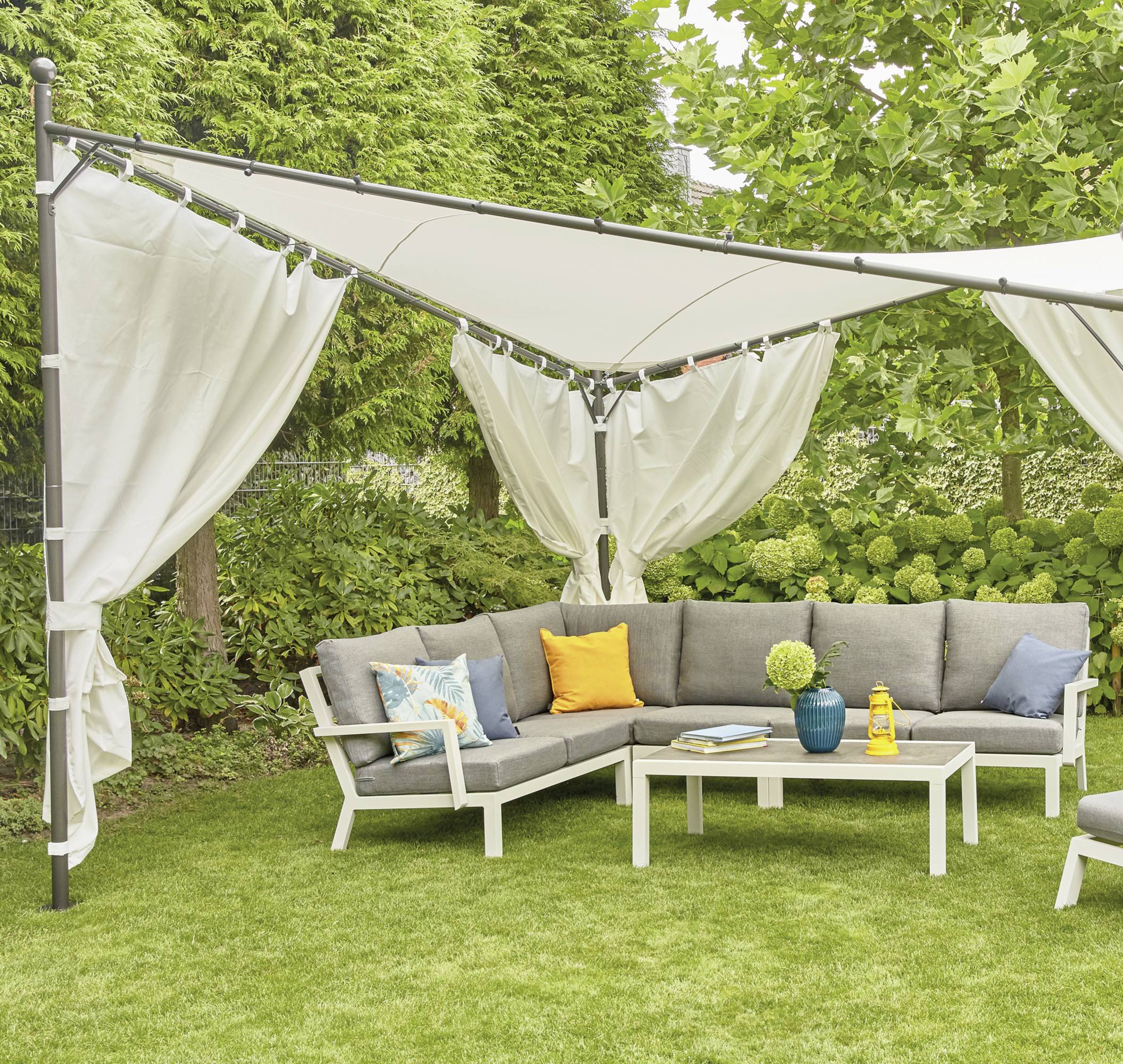 Sitzecke im Garten mit grauem Ecksofa, Tisch und Pavillon. Umgeben von grünen Bäumen und Pflanzen, ideal zum Entspannen im Freien.