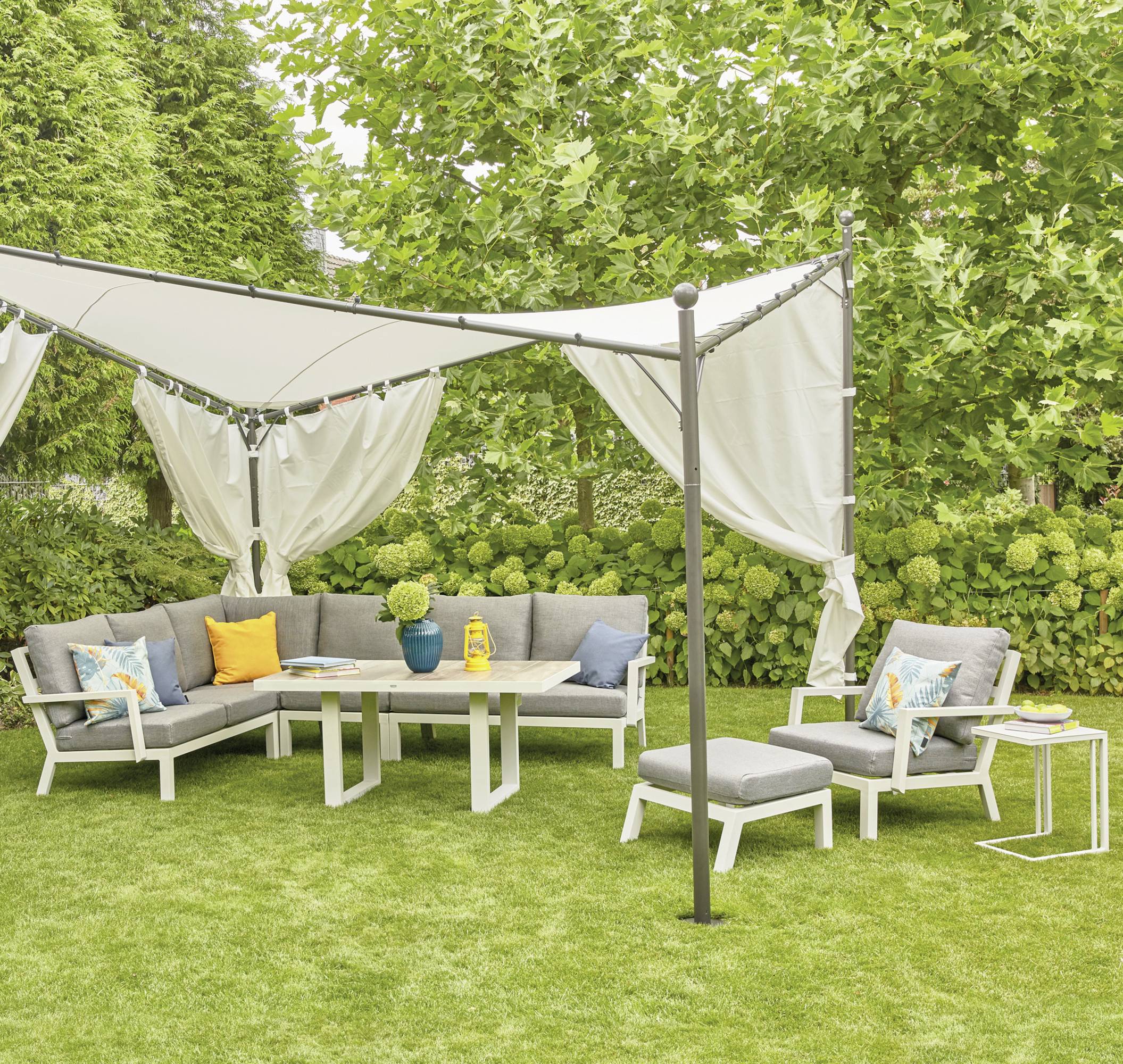 Garten mit grauen Sofas und einem Tisch auf grünem Rasen, umgeben von Bäumen. Im Hintergrund hängt eine weiße Stoffüberdachung.