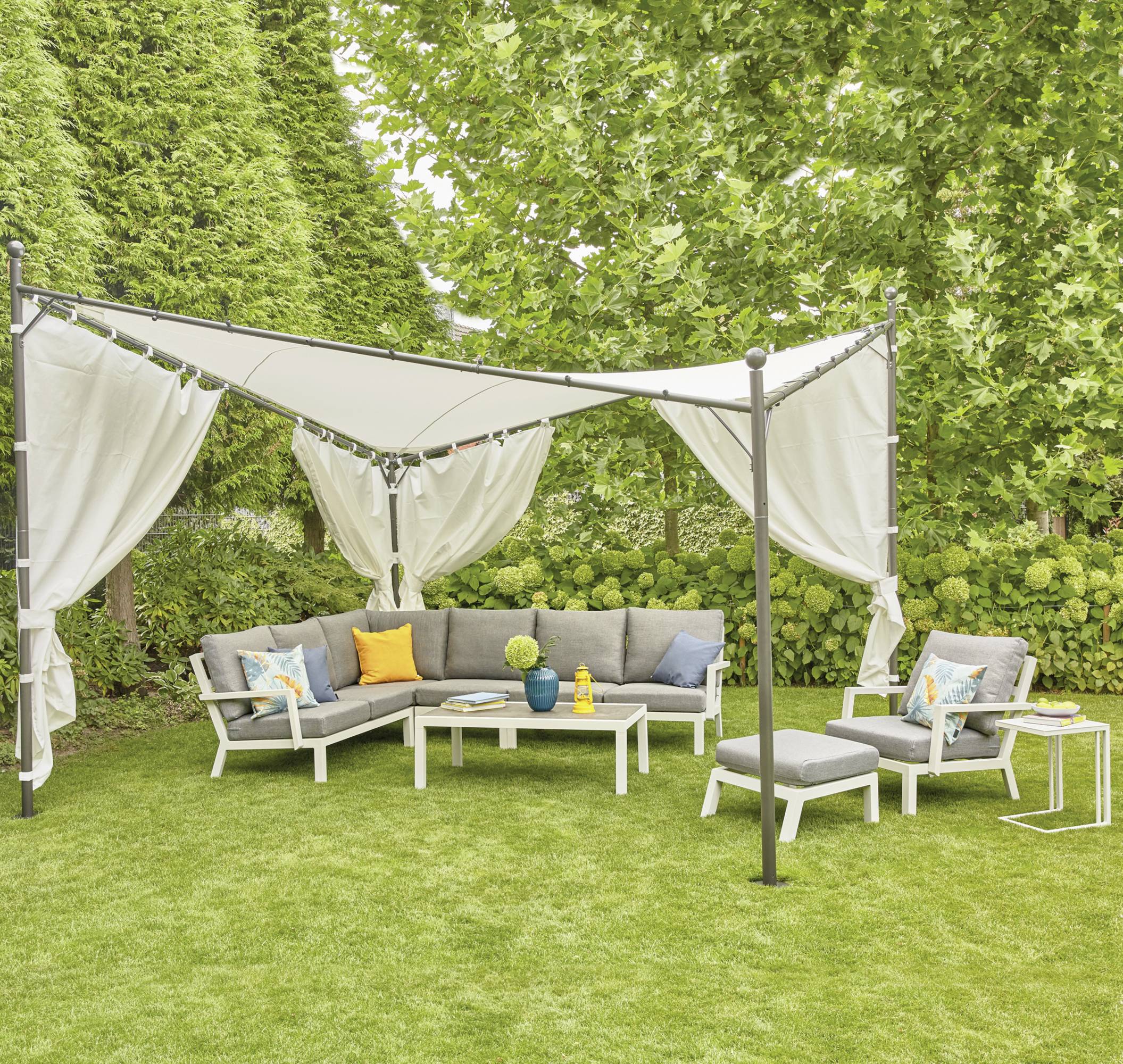 Ein Garten ist fotografiert: Ein gepolstertes Ecksofa mit bunten Kissen unter einem Sonnensegel auf einer grünen Rasenfläche.