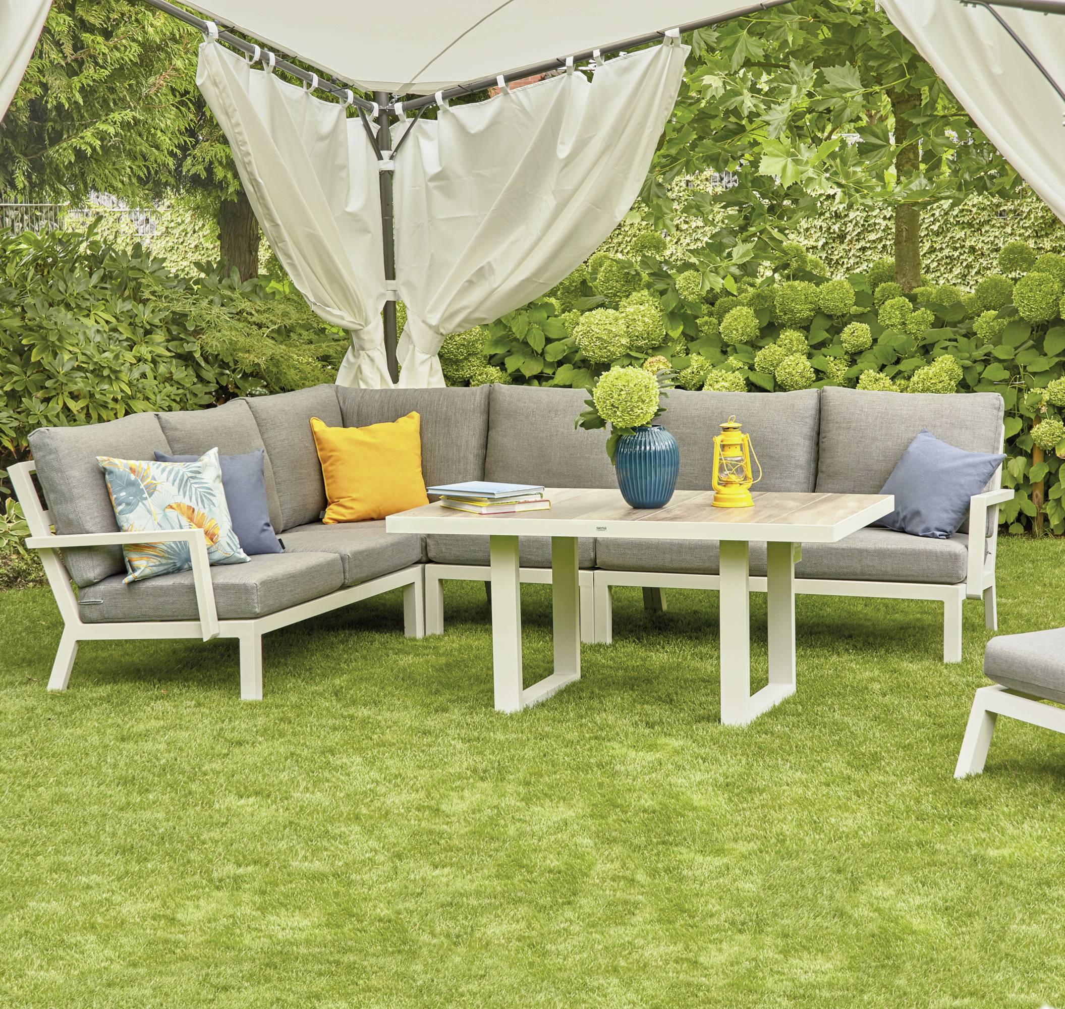 Gartenmöbel-Sitzgruppe mit grauen Kissen auf grünem Rasen unter einem Pavillon. Tisch in der Mitte mit Vase und Büchern.