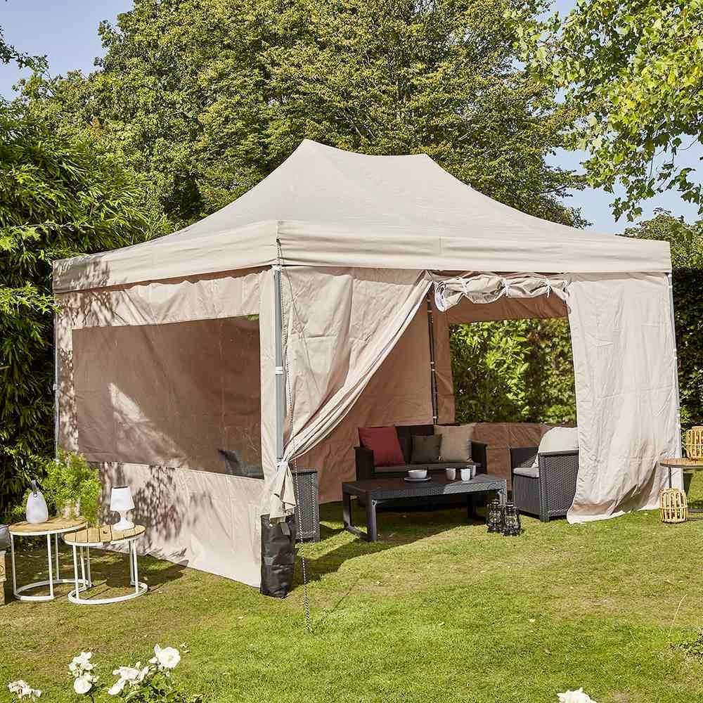 Ein beiger Pavillon steht im Garten auf grünem Rasen, umgeben von Bäumen. Innen befinden sich Gartenmöbel, darunter ein Sofa und ein Tisch.