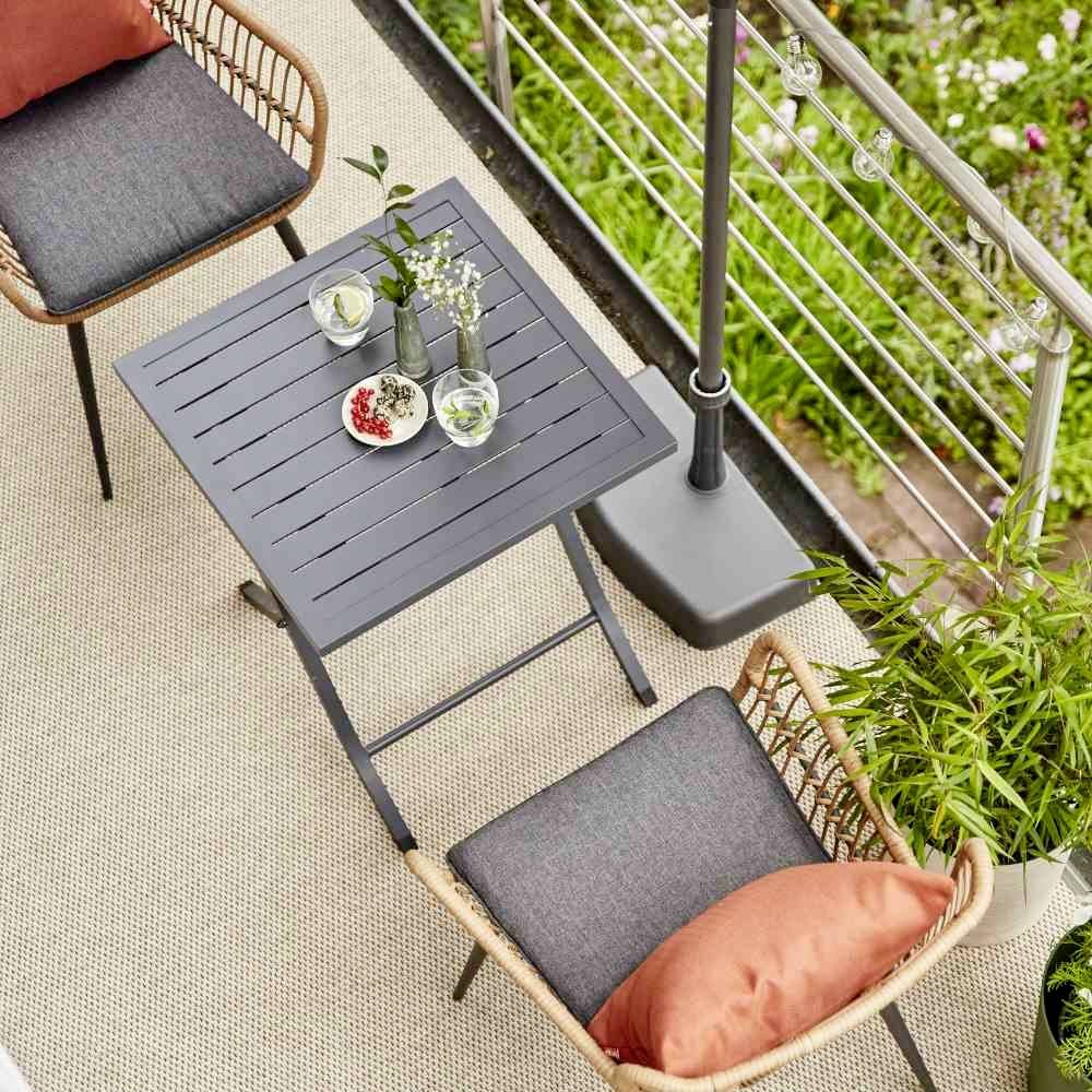 Zwei Stühle mit Kissen umgeben einen kleinen Tisch auf einem Balkon. Auf dem Tisch stehen Getränke und Blumen. Im Hintergrund Pflanzen.