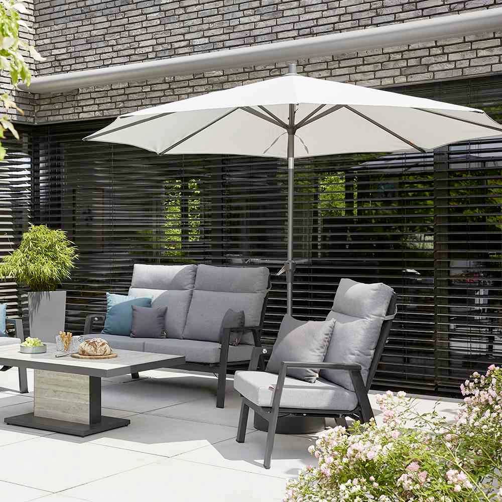 Terrasse mit grauen Sitzmöbeln, einem rechteckigen Tisch und einem weißen Sonnenschirm vor einem Gebäude mit großen Fenstern.