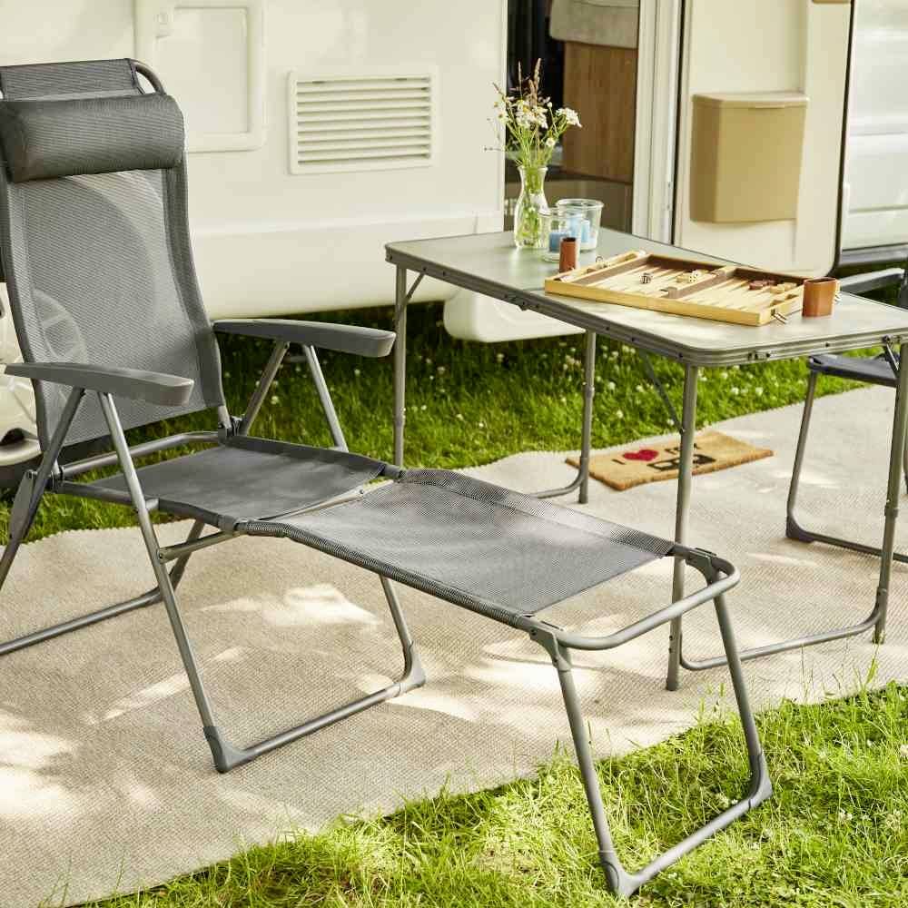 Gartenstuhl und Tisch mit Backgammon-Set, Vase und Getränken auf einem Teppich im Freien, umgeben von einem Wohnwagen.