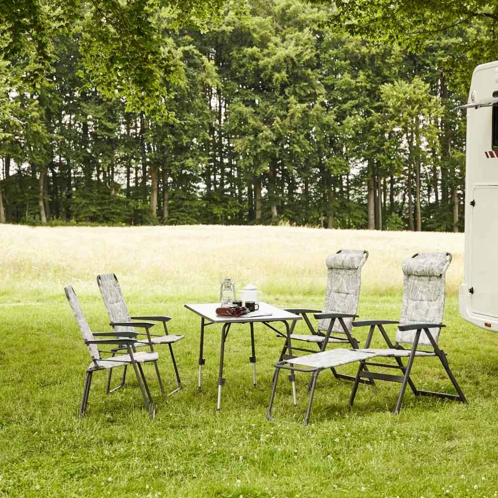 Ein Tisch und vier Stühle stehen auf einer Wiese vor einem Wohnmobil. Im Hintergrund sind Bäume. Camping-Szene in der Natur.
