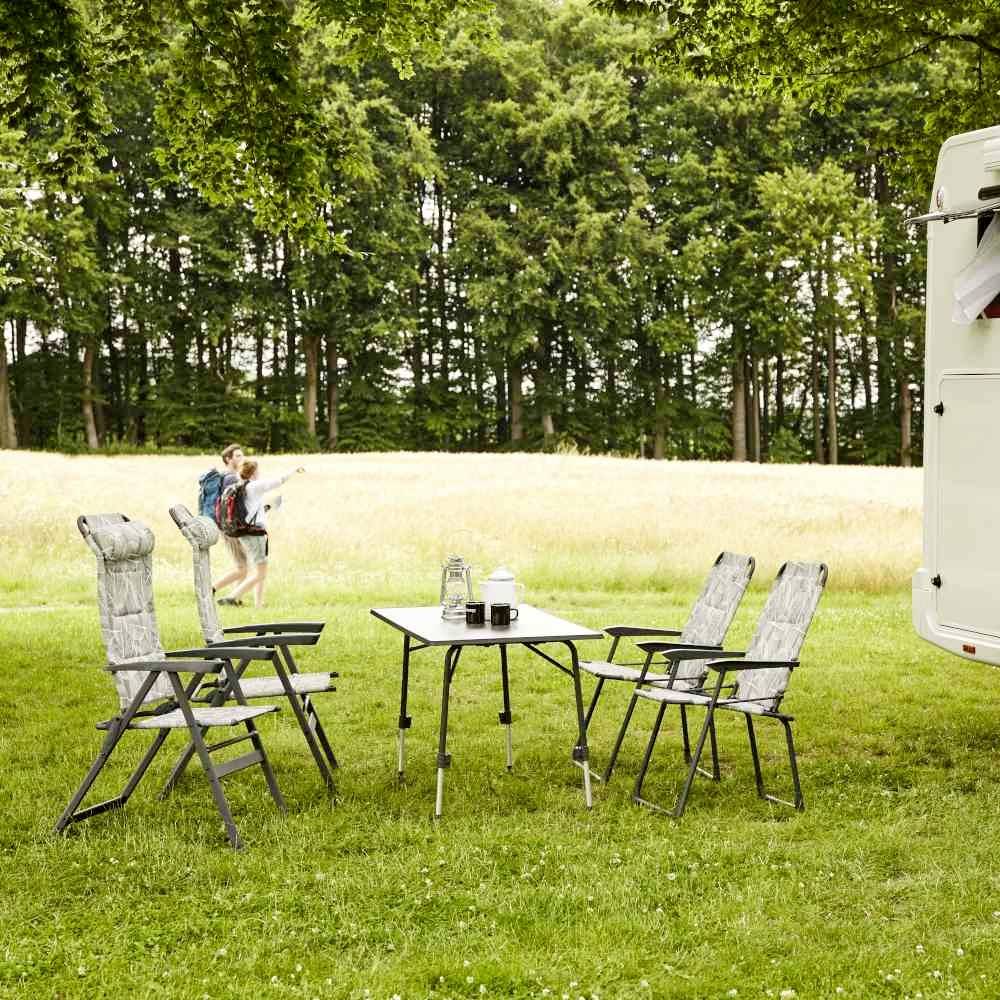 Campingplatz im Wald; zwei Personen mit Rucksäcken gehen im Hintergrund. Im Vordergrund steht ein Campingtisch mit vier Stühlen.