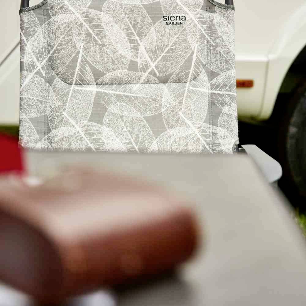 Gartenstuhl mit geblättertem Muster steht vor einem Wohnwagen. Im Vordergrund unscharfe Objekte. Der Stuhl wirkt einladend.