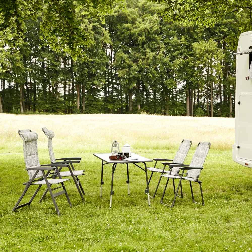 Ein Campingtisch mit vier Stühlen steht auf einer Wiese. Im Hintergrund sieht man einen Wohnwagen und Bäume. Ideal für Outdoor-Aktivitäten.
