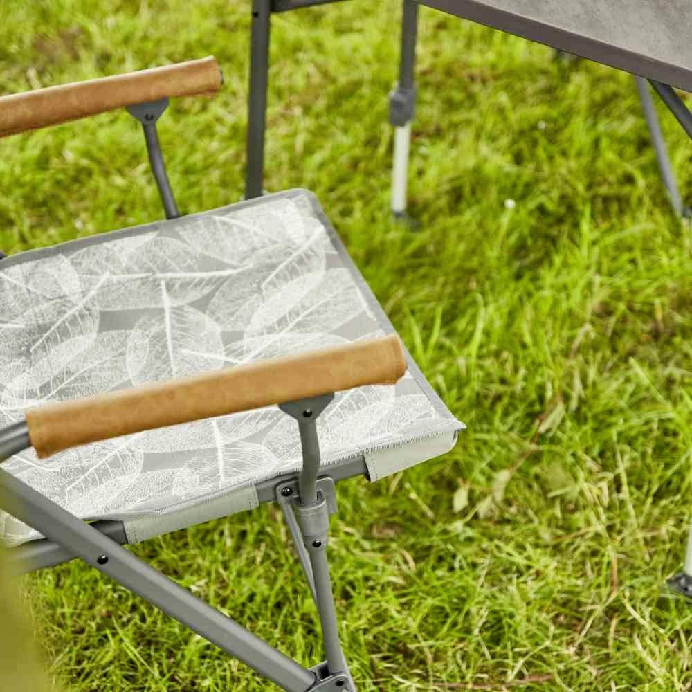 Ein Klappstuhl mit weißem, blattgemustertem Stoff sitzt auf einer grünen Wiese neben einem Tisch mit dunkler Oberfläche.
