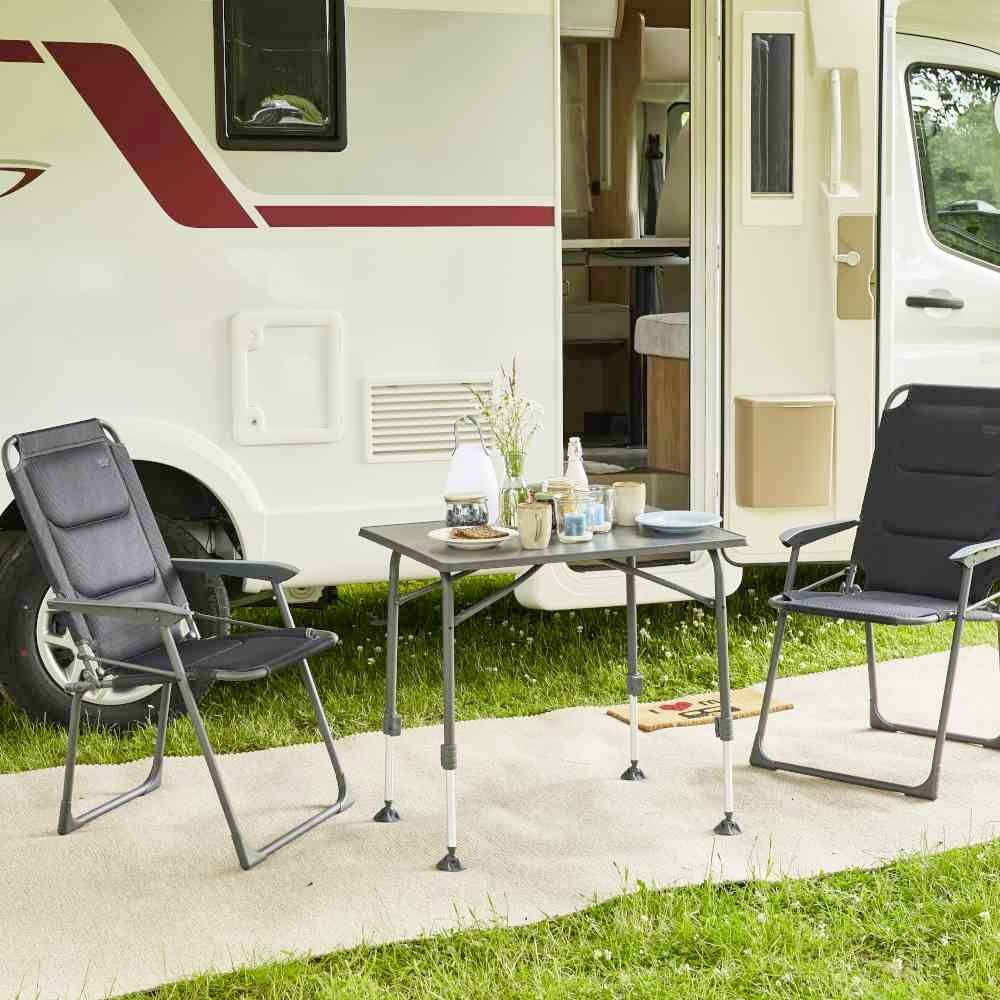 Wohnmobil mit geöffneter Tür auf einer Wiese. Davor stehen zwei gepolsterte Klappstühle und ein Tisch mit Vase und Tassen.