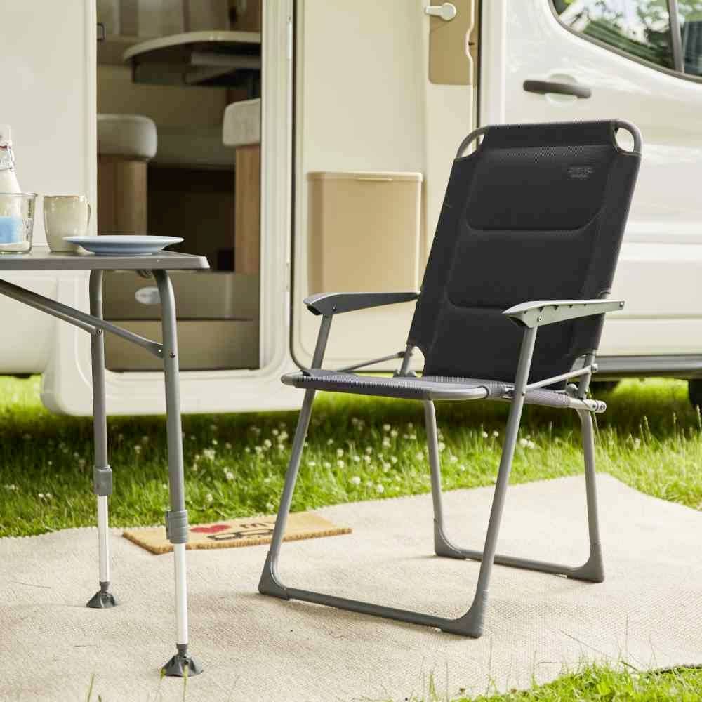 Ein Klappstuhl und ein Tisch mit Geschirr stehen auf einem Teppich vor einem Wohnwagen, der auf einer grünen Wiese geparkt ist.