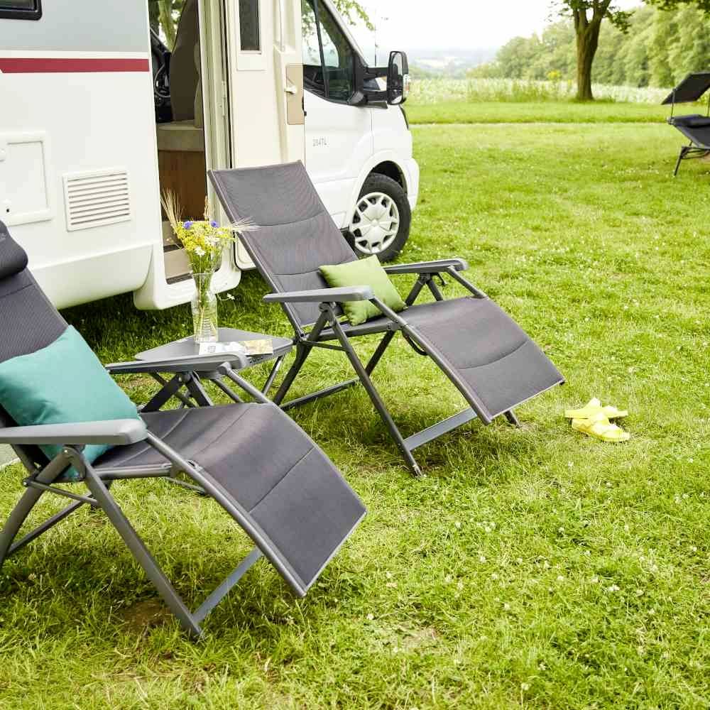 Wohnmobil auf einer Wiese mit geöffnetem Eingang. Zwei Sonnenliegen und ein Tisch stehen daneben, Vase mit Blumen darauf. Ruhige ländliche Kulisse.