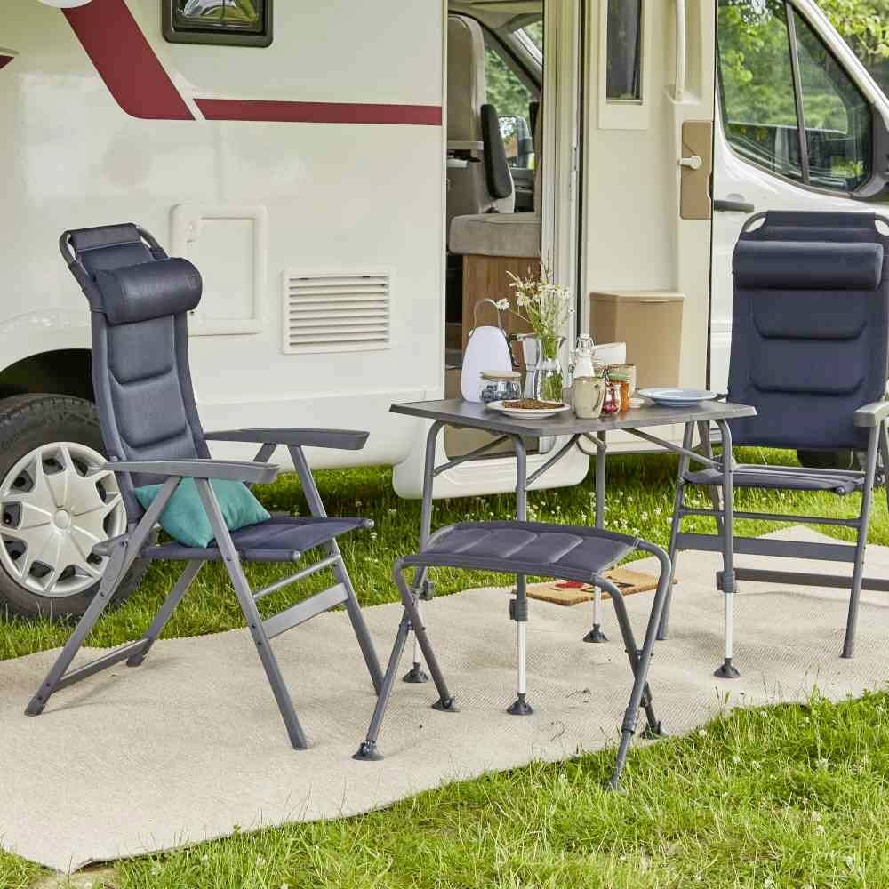 Wohnwagen mit offenem Innenraum, umgeben von zwei Campingstühlen und einem Tisch mit Geschirr, auf grünem Rasen platziert.