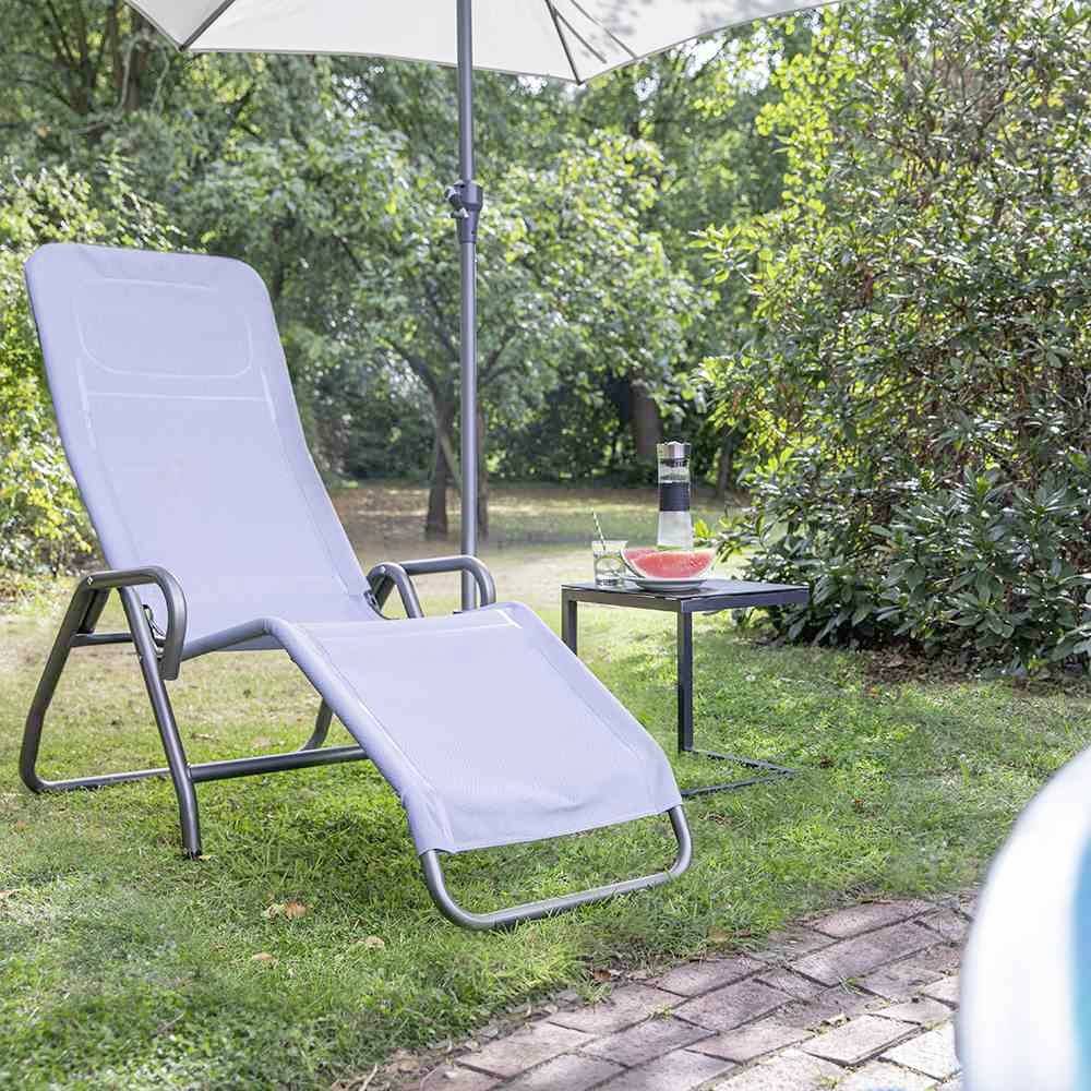 Ein Liegestuhl im Garten steht unter einem Sonnenschirm. Daneben ein kleiner Tisch mit einer Karaffe und Schale mit Melone. Grüne Büsche im Hintergrund.