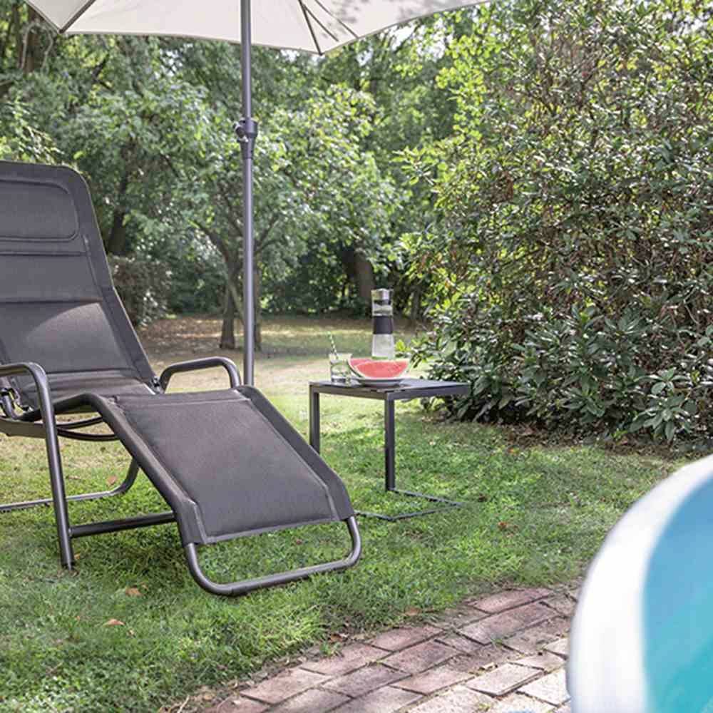 Ein Liegestuhl und Tisch mit Wasserflasche und Schale unter einem Sonnenschirm, im Grünen mit Bäumen und Sträuchern im Hintergrund.
