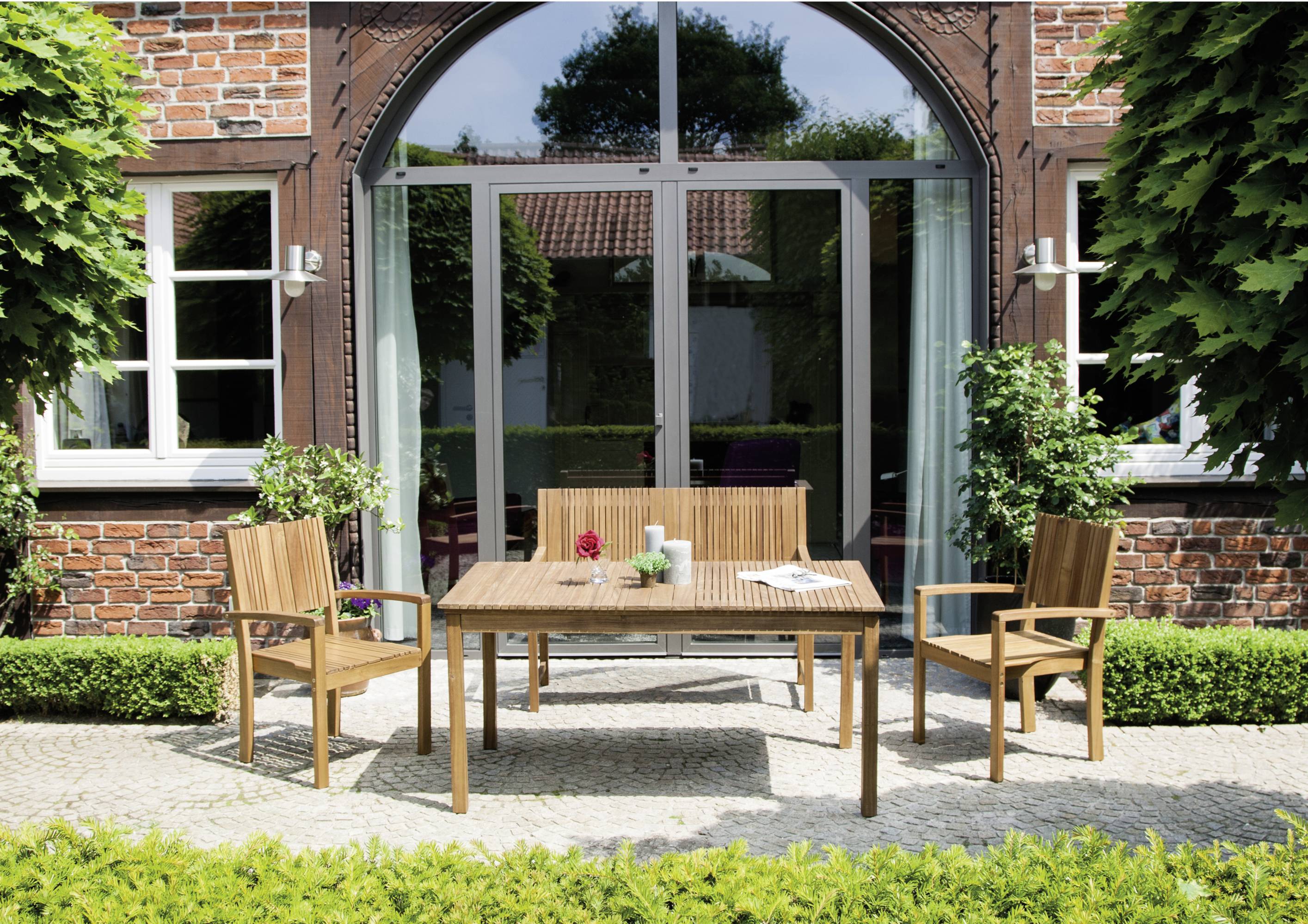Holztisch und Stühle auf einer Terrasse vor einem Backsteinhaus mit großen Fenstern und grüner Hecke im Hintergrund.