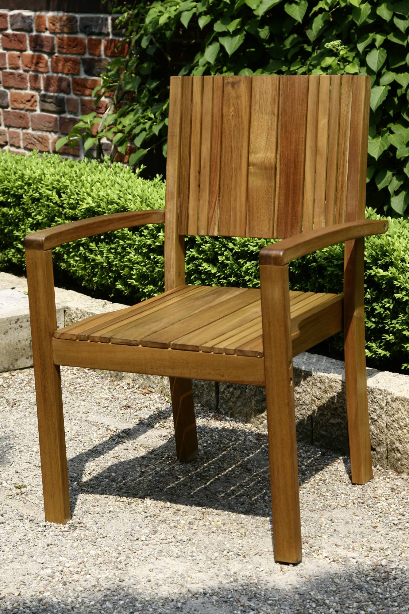 Holzstuhl mit Armlehnen steht auf einem Kiesboden im Garten, umgeben von grünen Büschen und einer Backsteinmauer im Hintergrund.