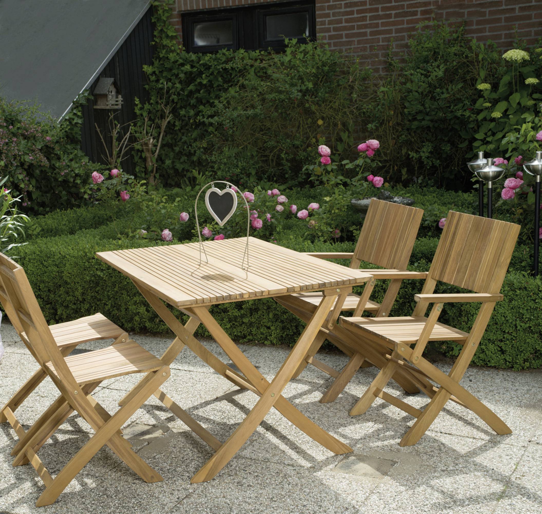 Holzgarnitur auf Terrasse mit vier Stühlen und Tisch, umgeben von grünen Büschen und blühenden Rosen.