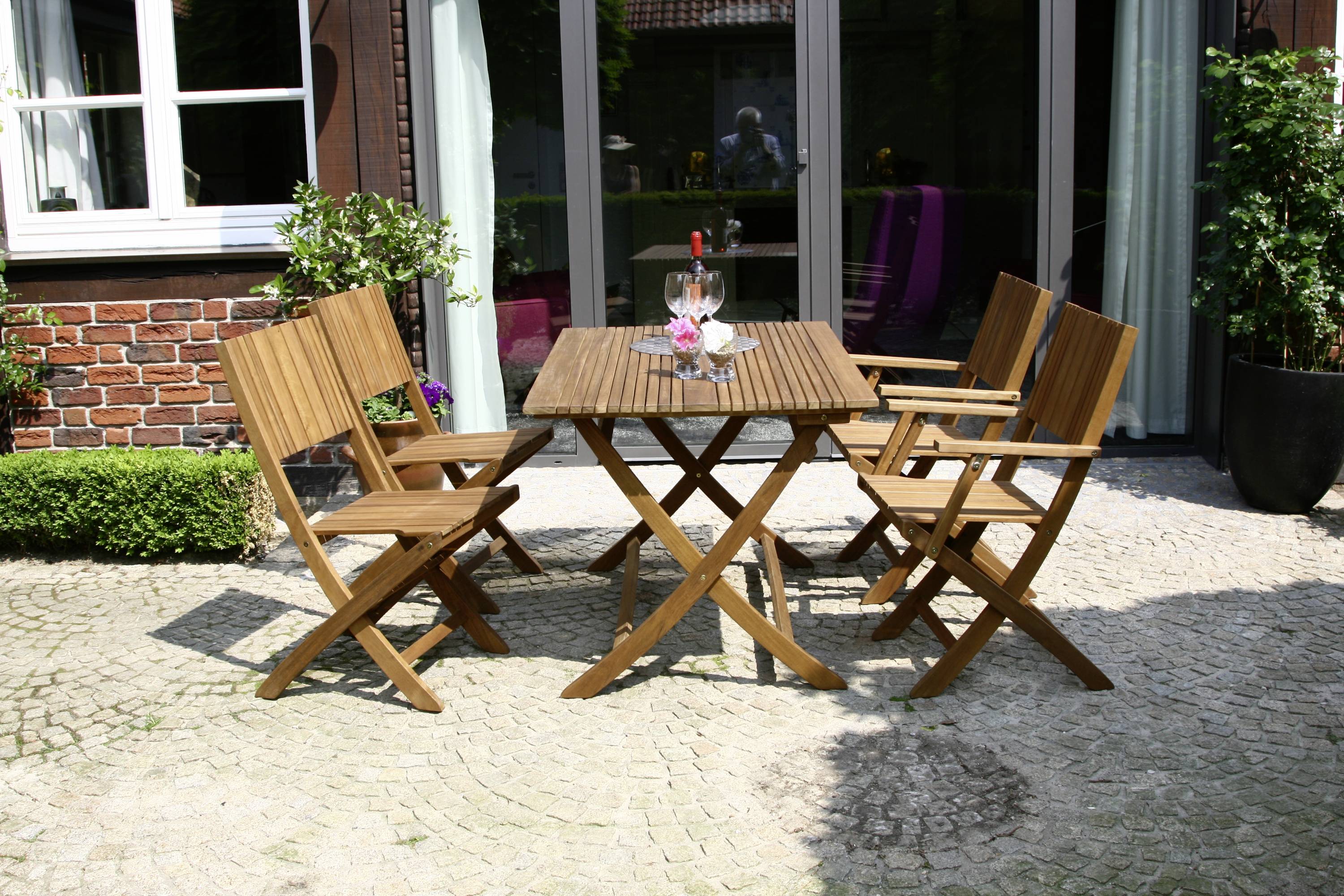 Ein Holztisch mit vier Stühlen steht auf einer gepflasterten Terrasse vor einer Hauswand. Auf dem Tisch befinden sich zwei Weingläser.